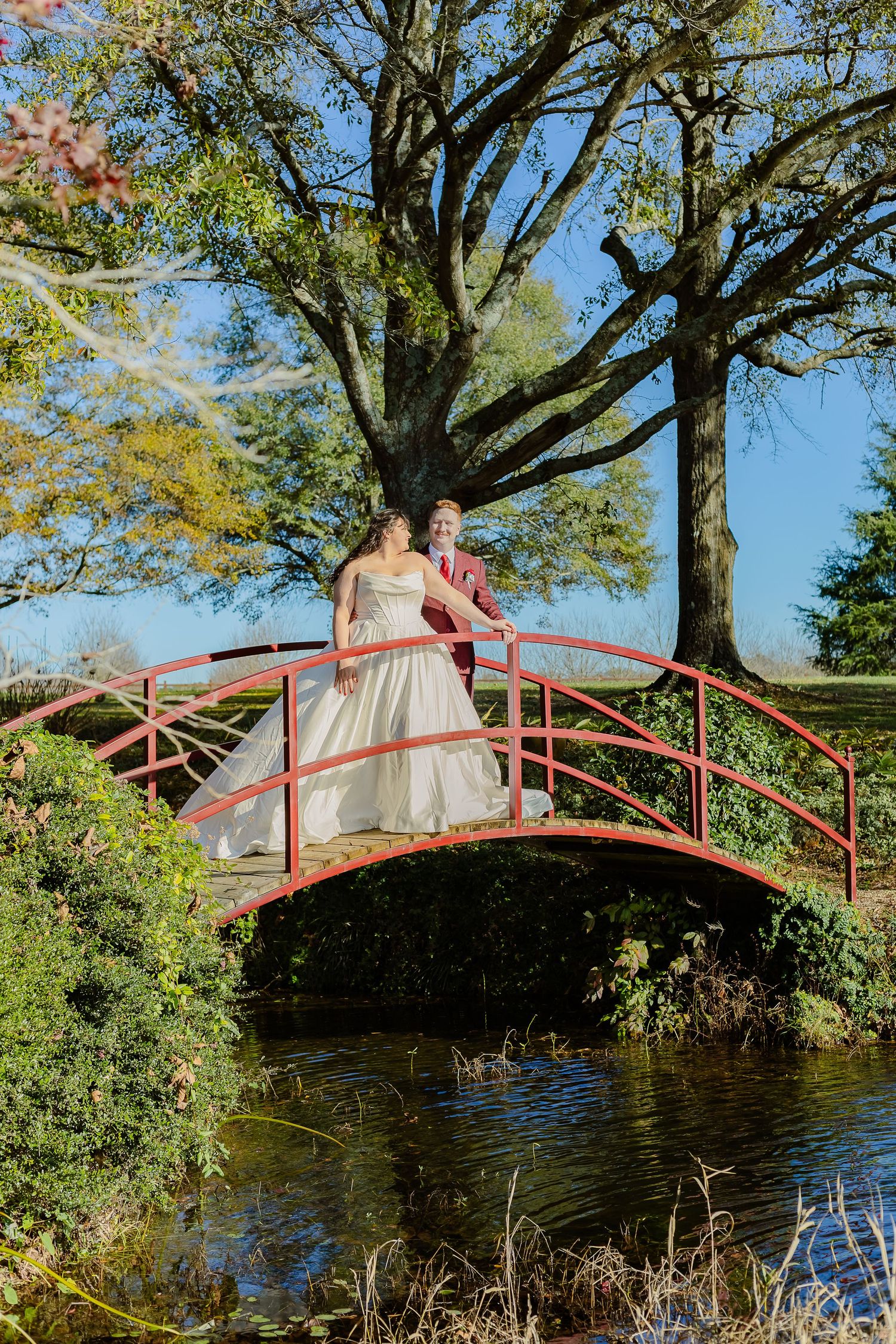 A couple in wedding attire stands on a red arched bridge over a pond surrounded by lush green trees on a sunny day.