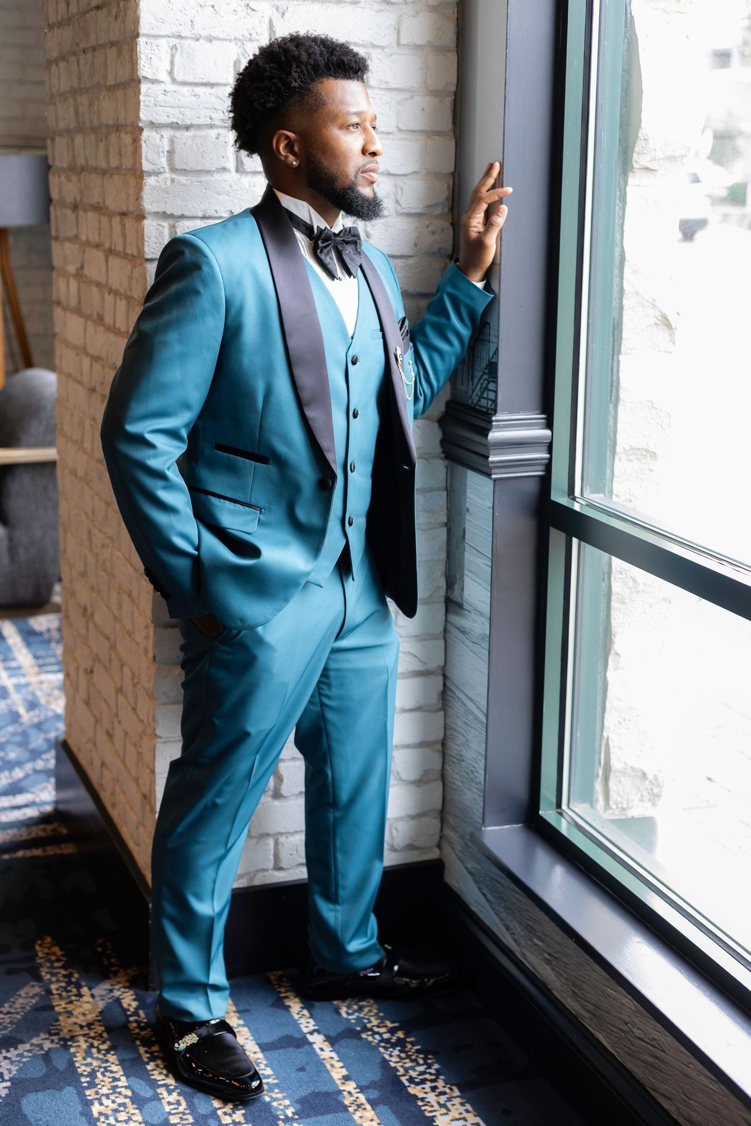 A stylish teal three-piece suit photographed against a brick wall near a window.