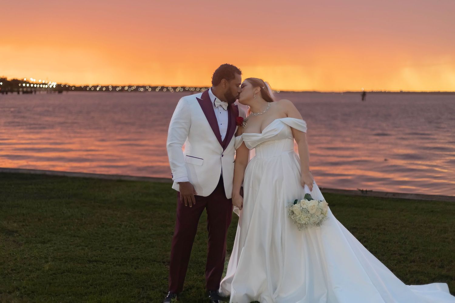 A wedding couple poses against a stunning orange and pink sunset on a beach with palm trees in the background.