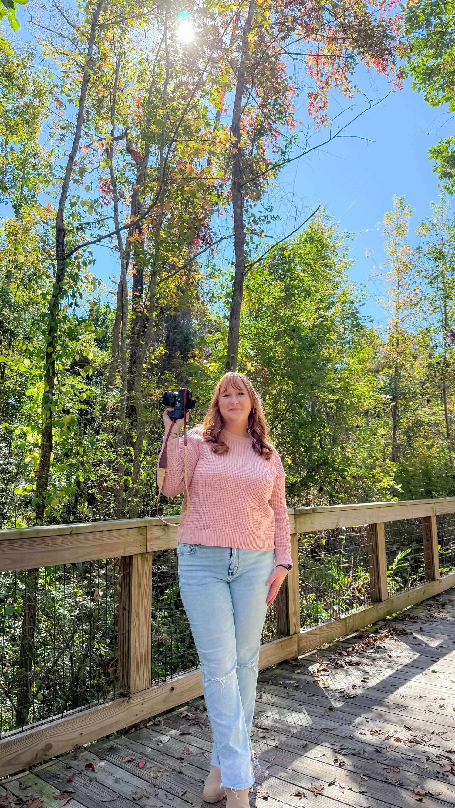 Person in pink sweater and jeans walking along a wooden boardwalk surrounded by autumn trees on a sunny day.