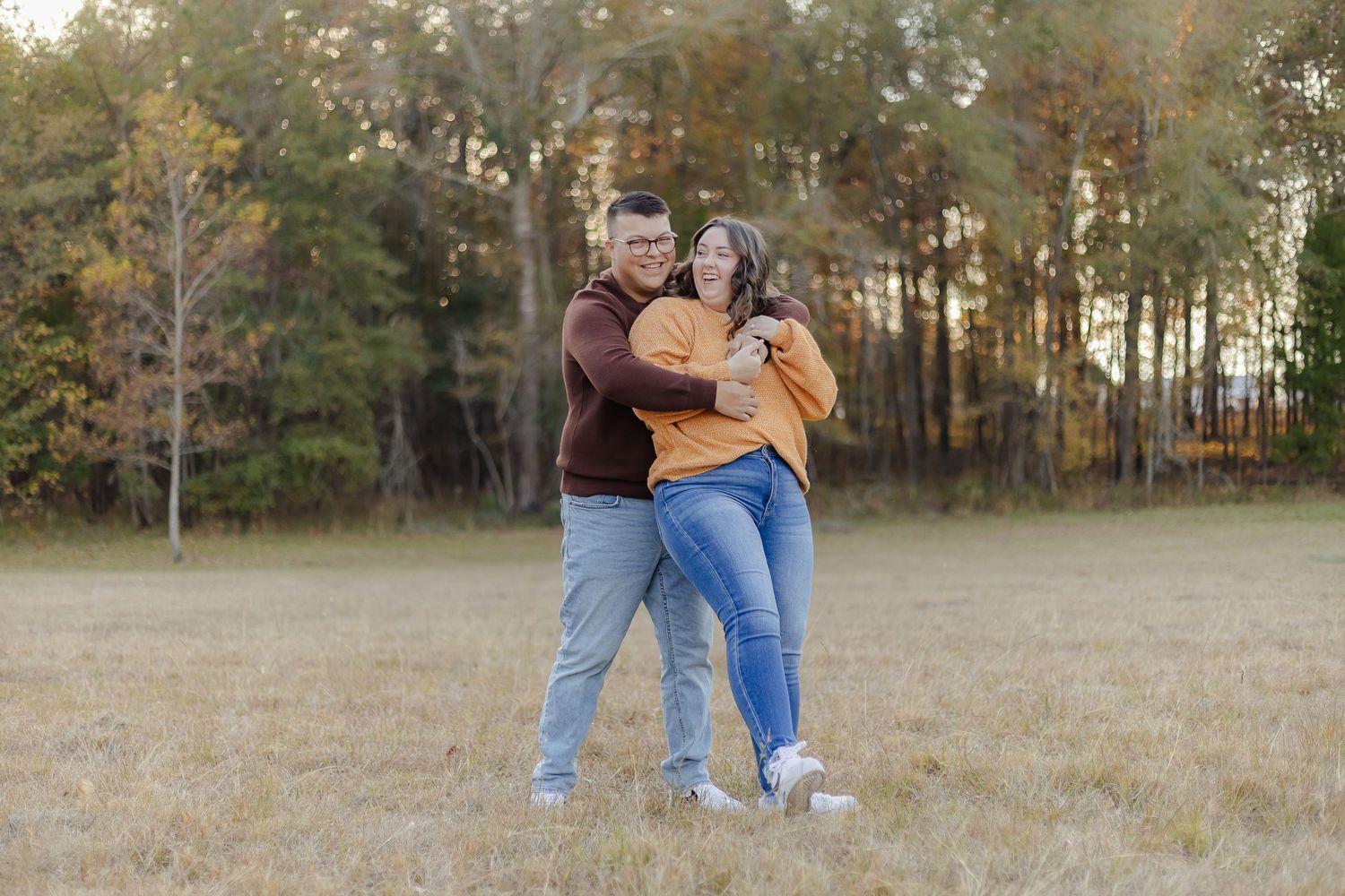 A couple embraces while standing in a golden autumn field with trees in the background during sunset.