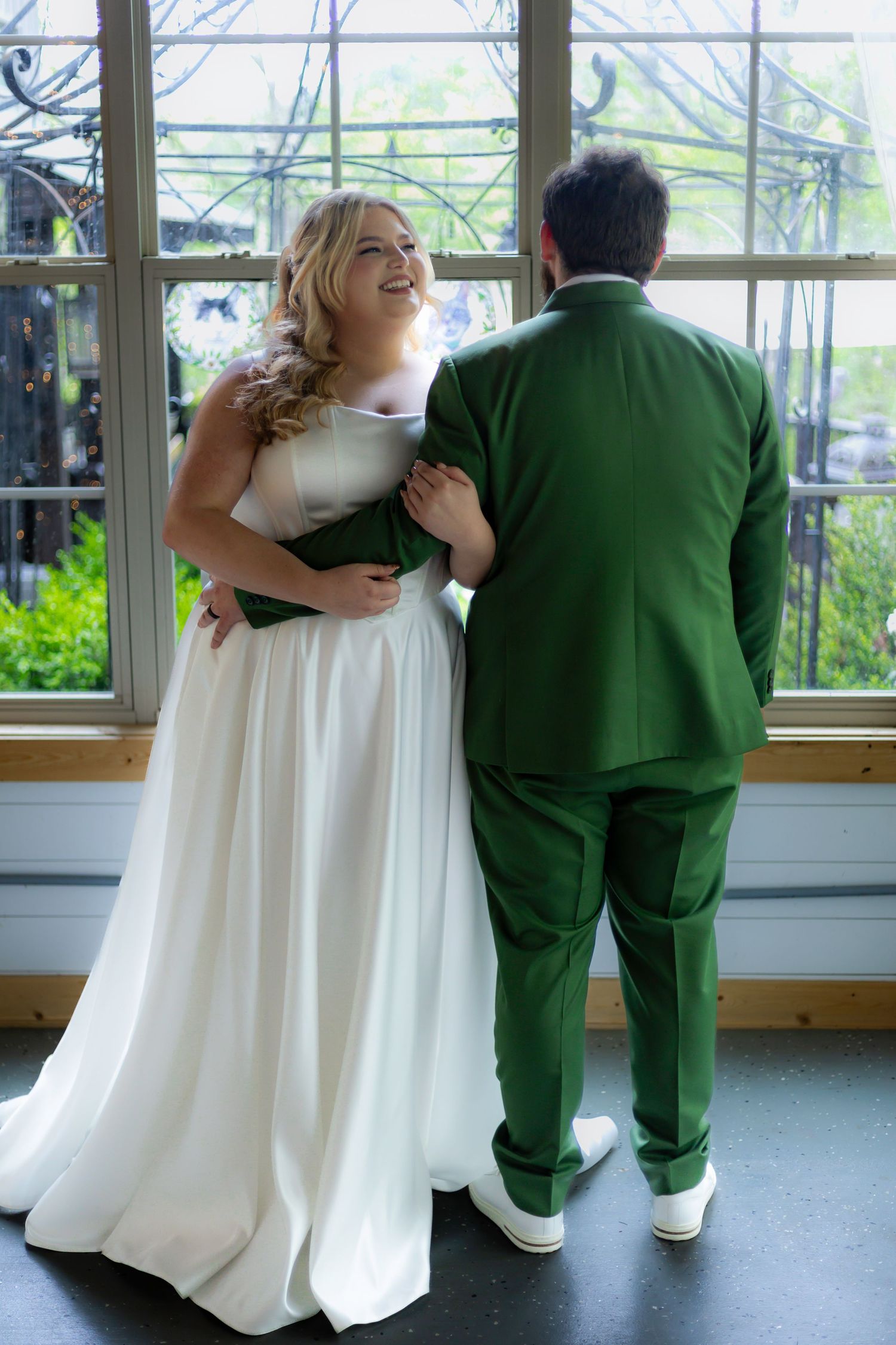 Wedding couple in white dress and striking green suit laughing by greenhouse windows.