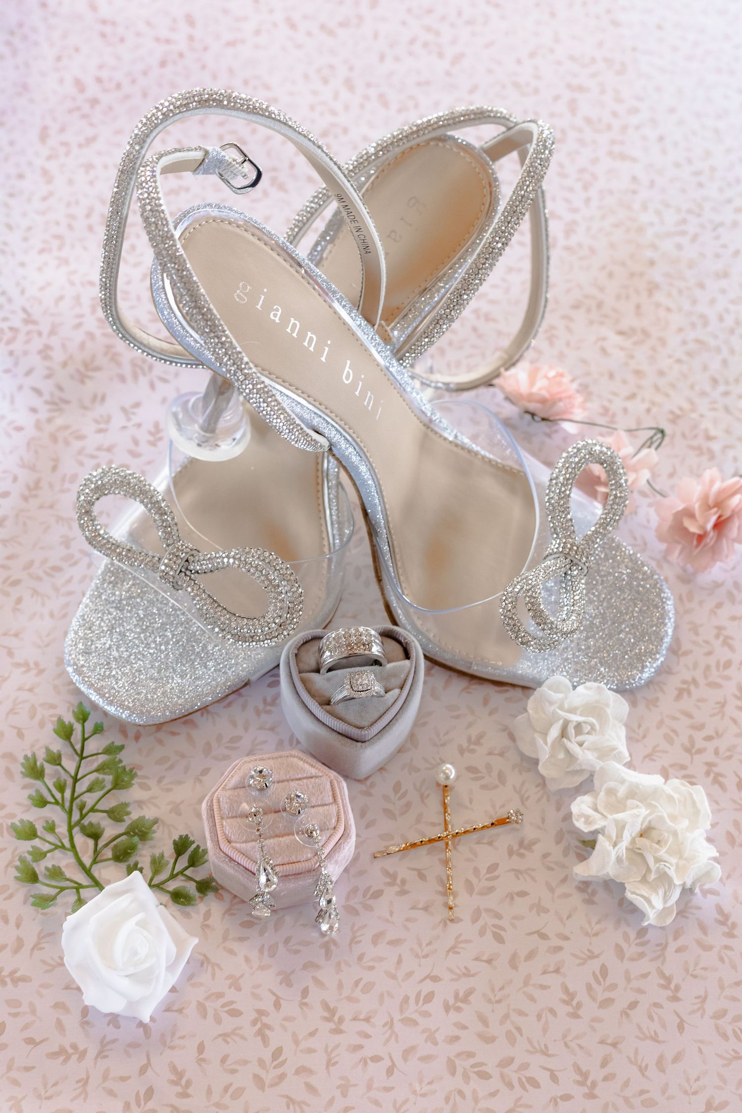 Sparkly silver bridal shoes with ankle straps styled with delicate white roses and pink floral accents on lace background.
