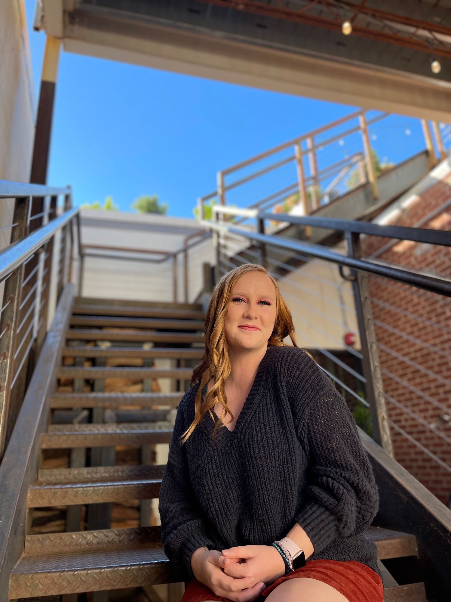 Person in black sweater posing on outdoor wooden stairs against brick building.