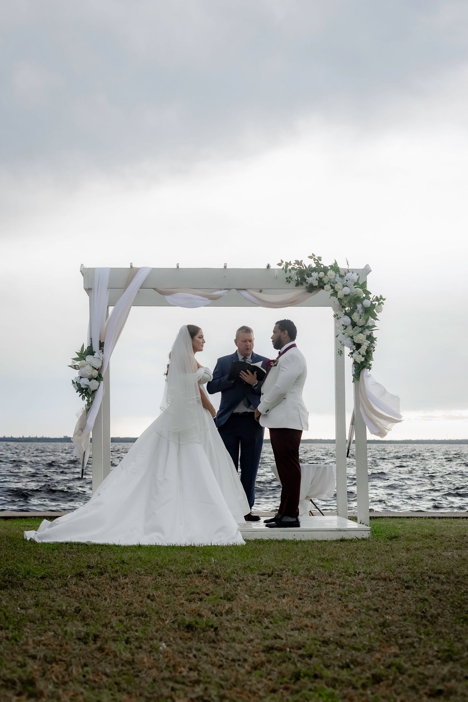 A wedding ceremony takes place under a white arbor on a waterfront lawn with cloudy skies in the background.