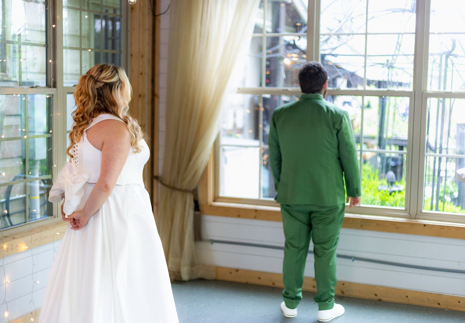 A couple in green and white wedding attire stands in front of large windows in a bright indoor venue.