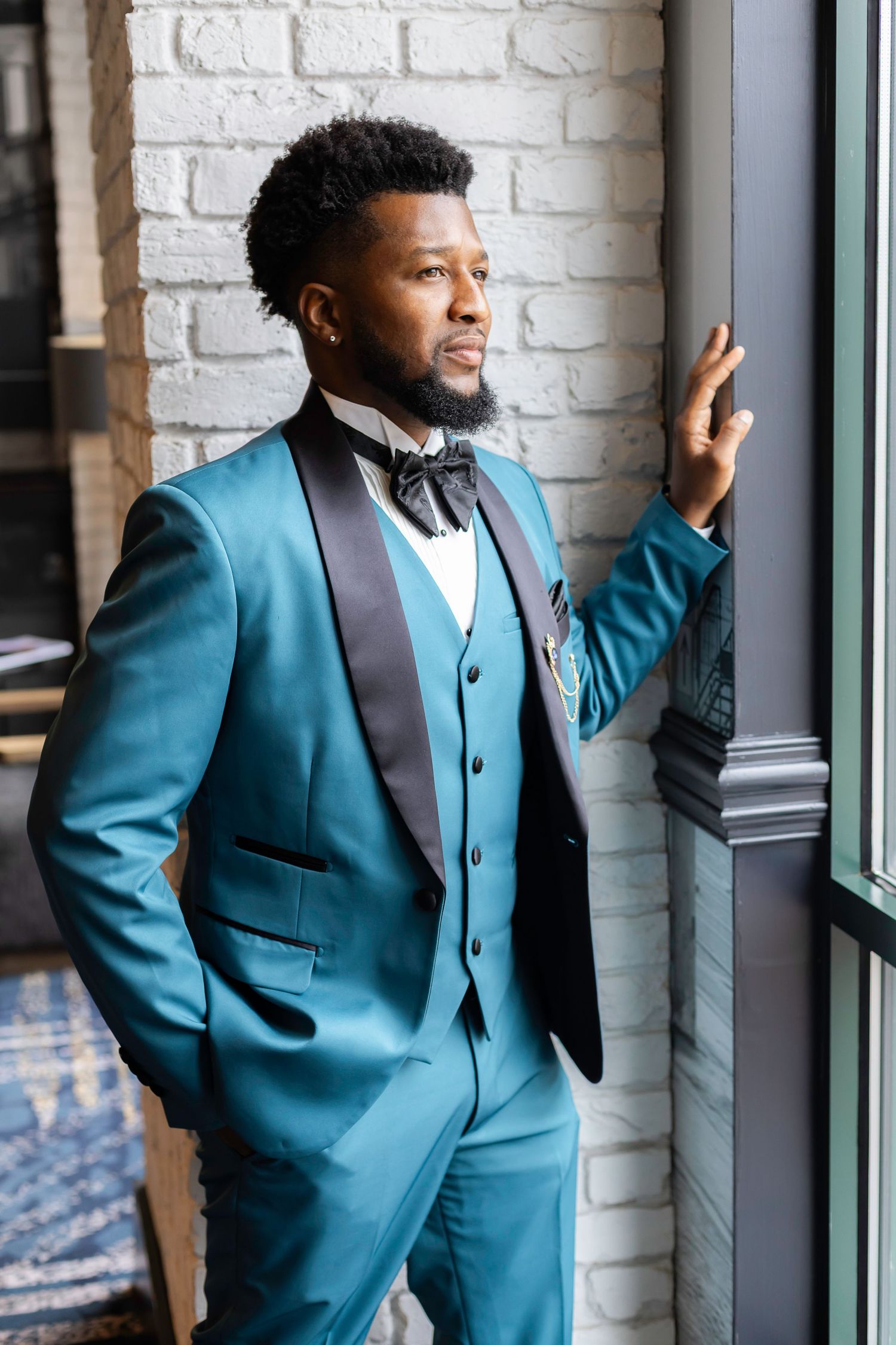 A groom poses in a stylish teal suit with black lapels against a white brick wall.