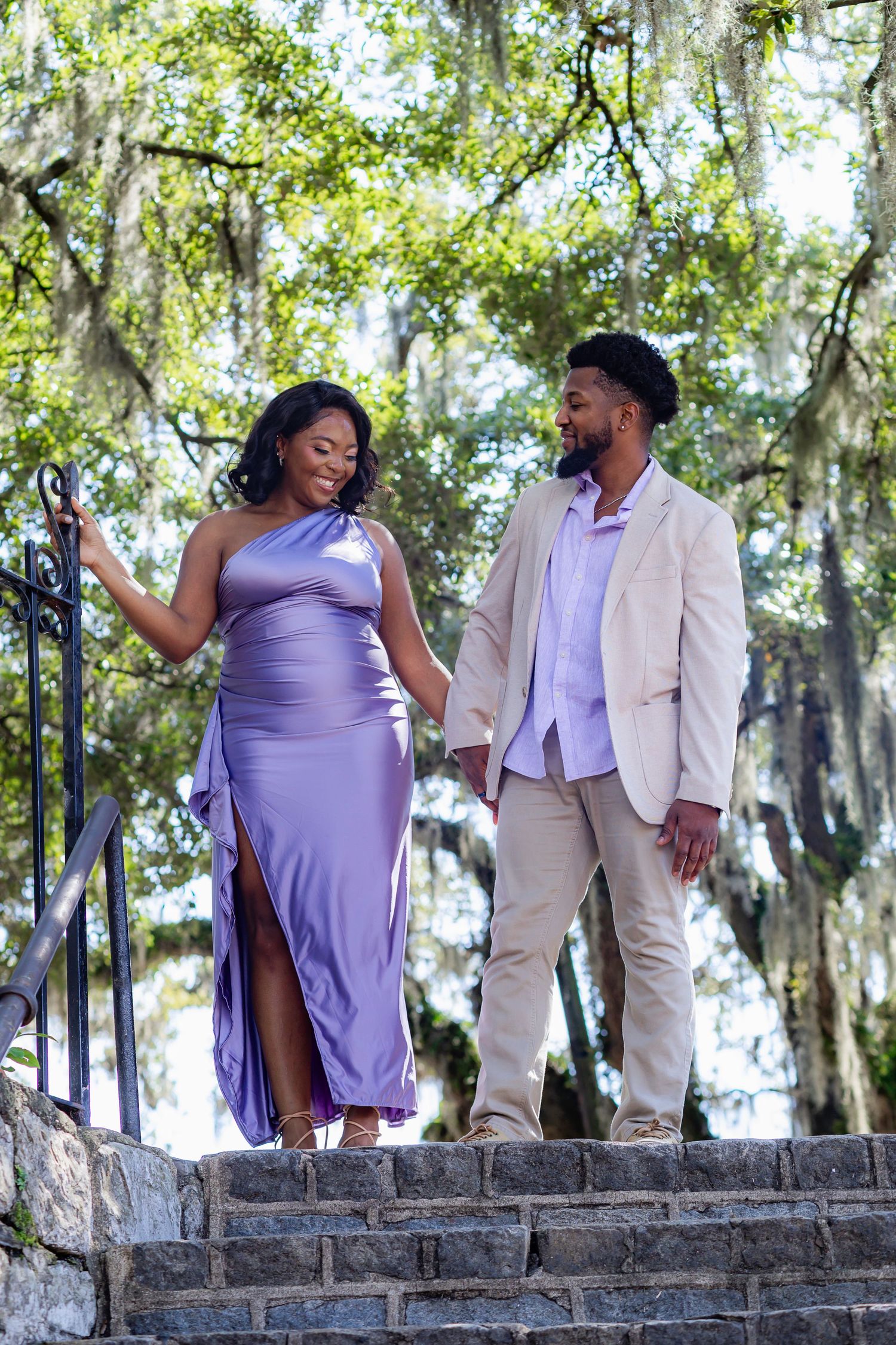 Elegant couple in formal attire standing on stone steps surrounded by Spanish moss and trees.