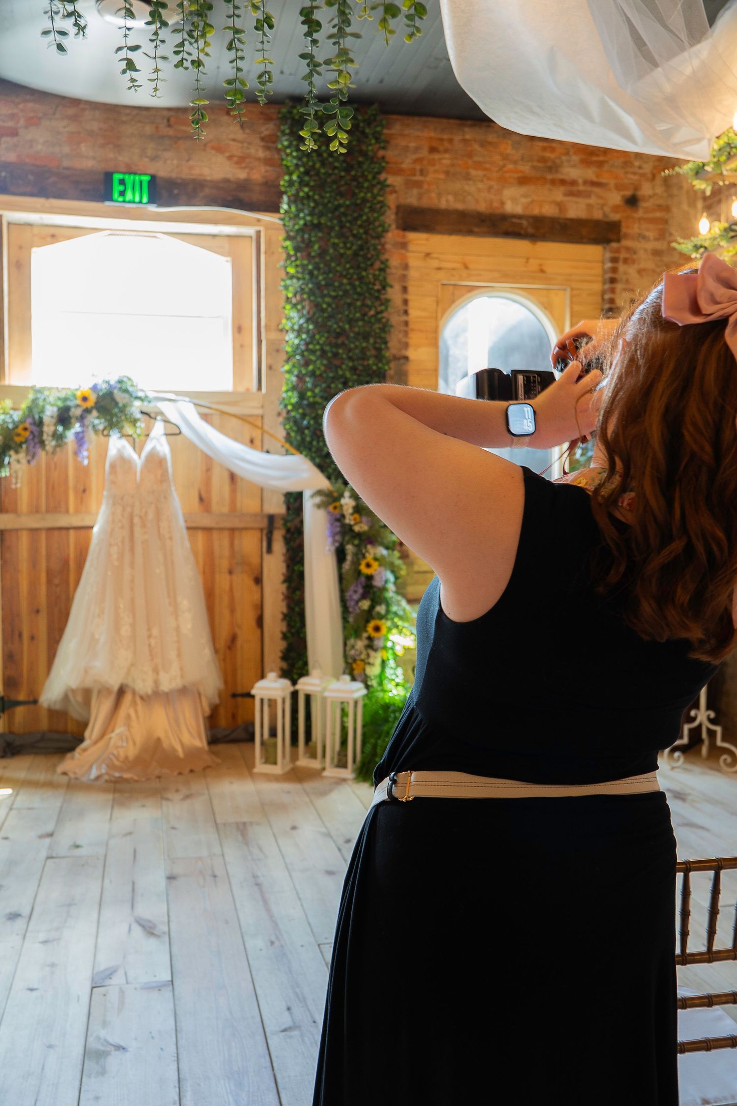 A wedding dress is displayed on a mannequin in a rustic brick-walled venue with greenery and white decor.