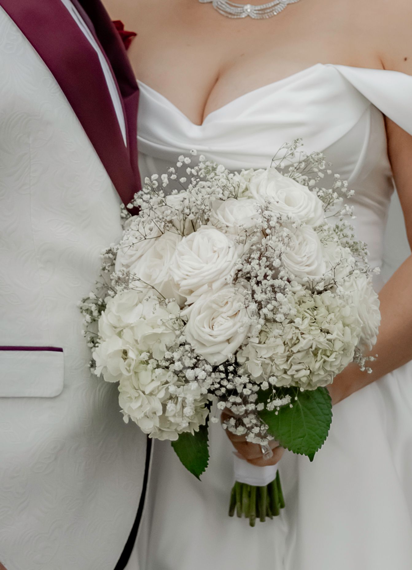 A white bridal bouquet featuring roses and baby's breath with burgundy ribbon wrapped around the stems.