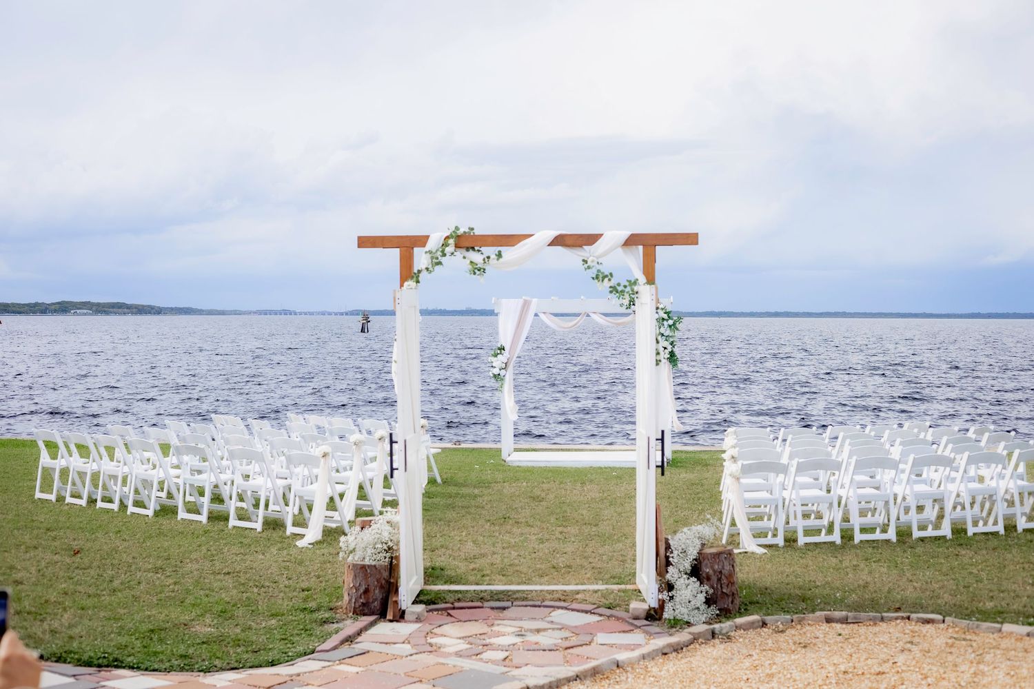 White wedding ceremony setup with wooden arch and chairs overlooking the ocean on a grassy waterfront lawn.