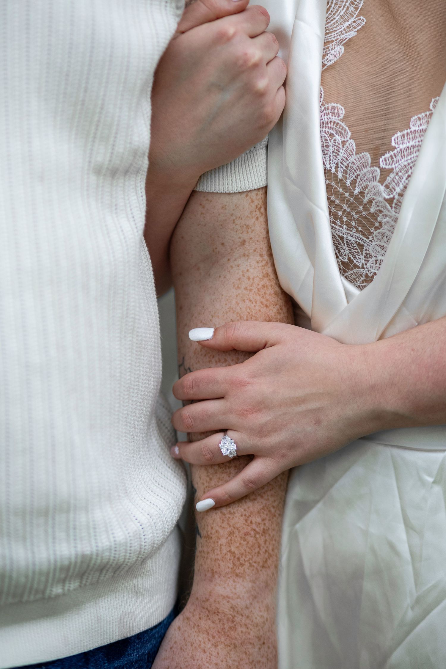 Close-up of hands embracing with white lace robe detail and engagement ring visible.