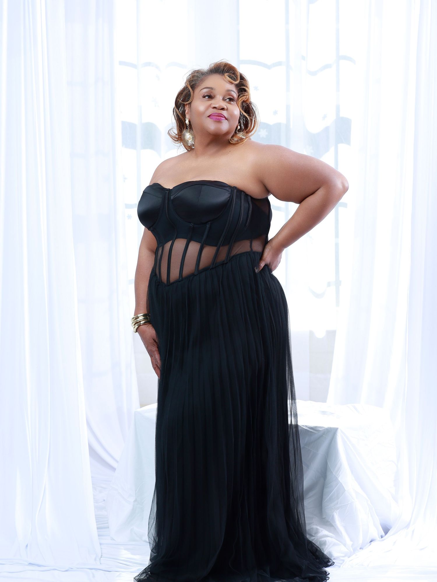 Woman in elegant black strapless corset gown with sheer pleated skirt posing confidently against white draped backdrop.