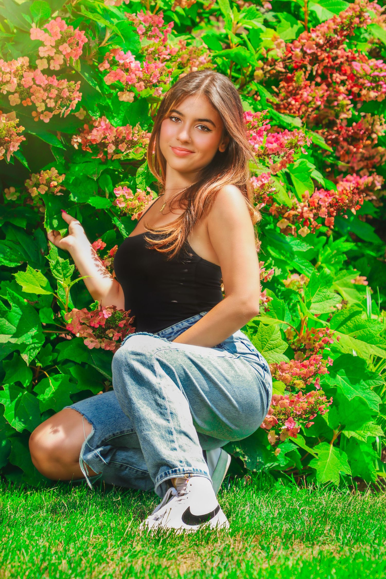 Woman in casual wear poses with pink flowering shrubs in vibrant garden setting.