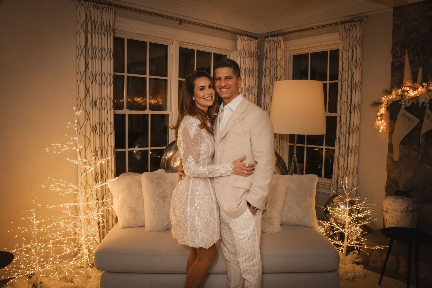 Couple in matching white outfits posing together in a warmly lit room decorated with glowing fairy light trees.
