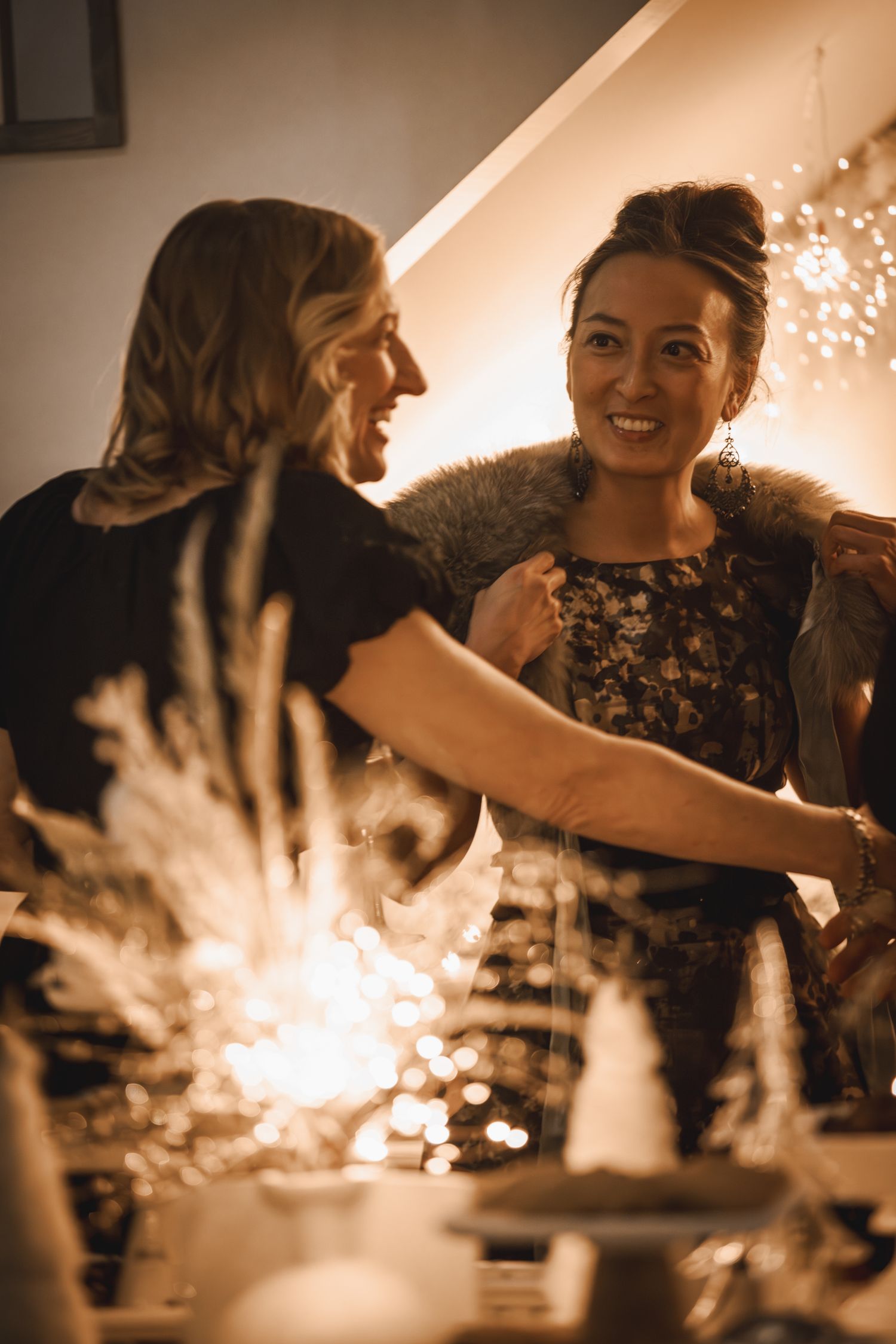 Two women smiling and laughing together at a festive holiday party with warm glowing lights in background.