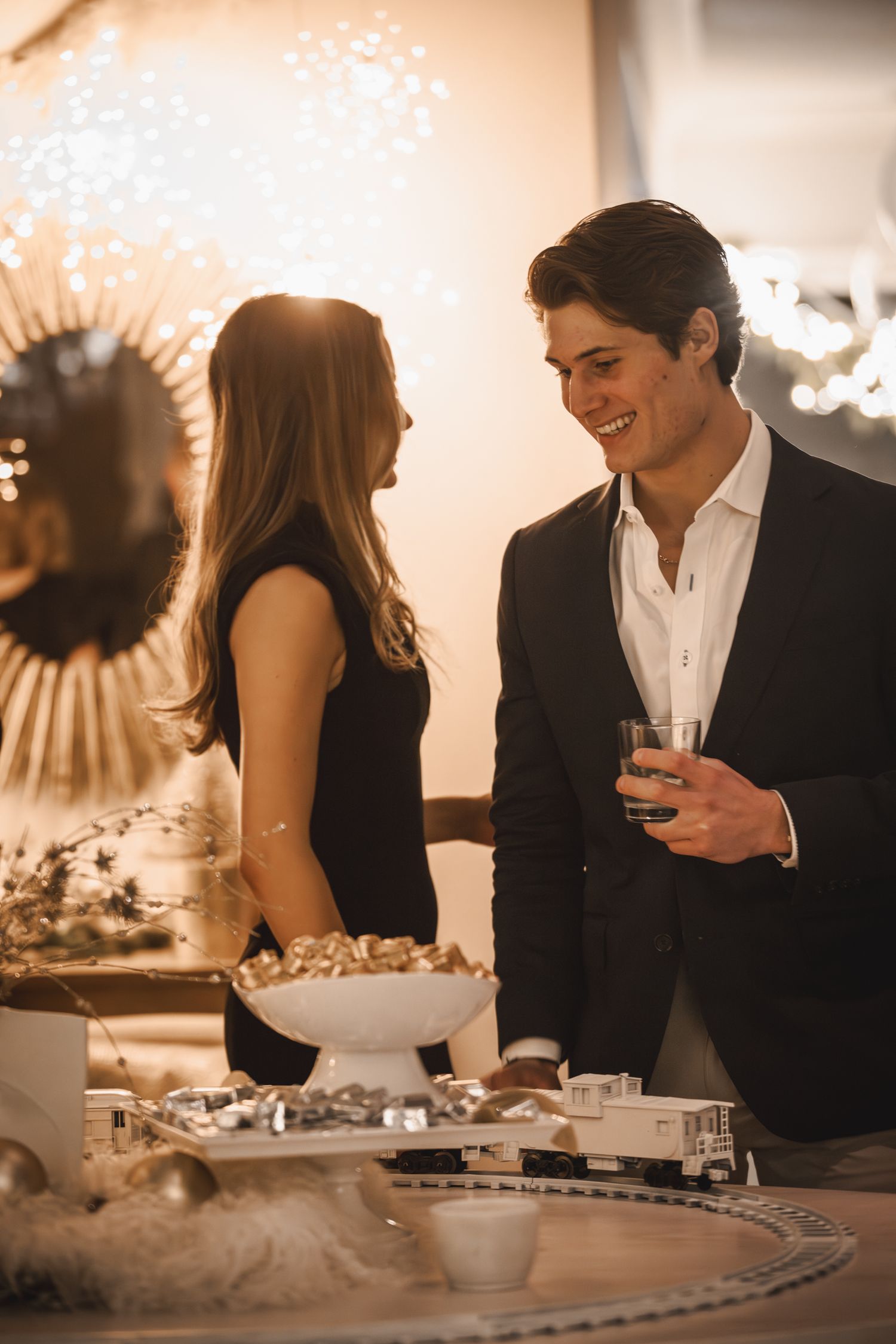 A smiling young man in a suit holds a drink while chatting with a woman at an elegant holiday party.