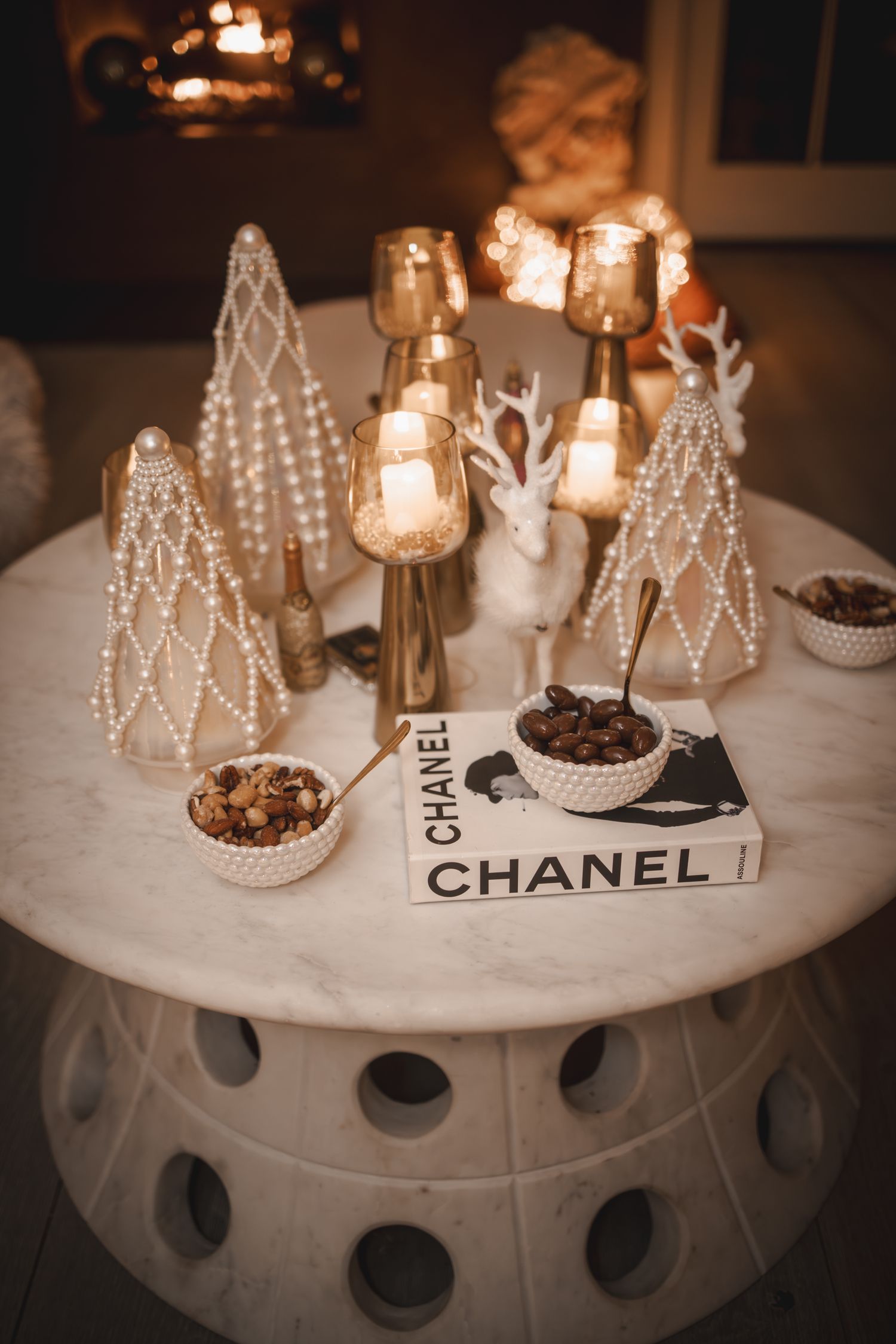 Elegant Christmas table with Chanel book, pearl trees, gold candles, and white reindeer decor on marble table.