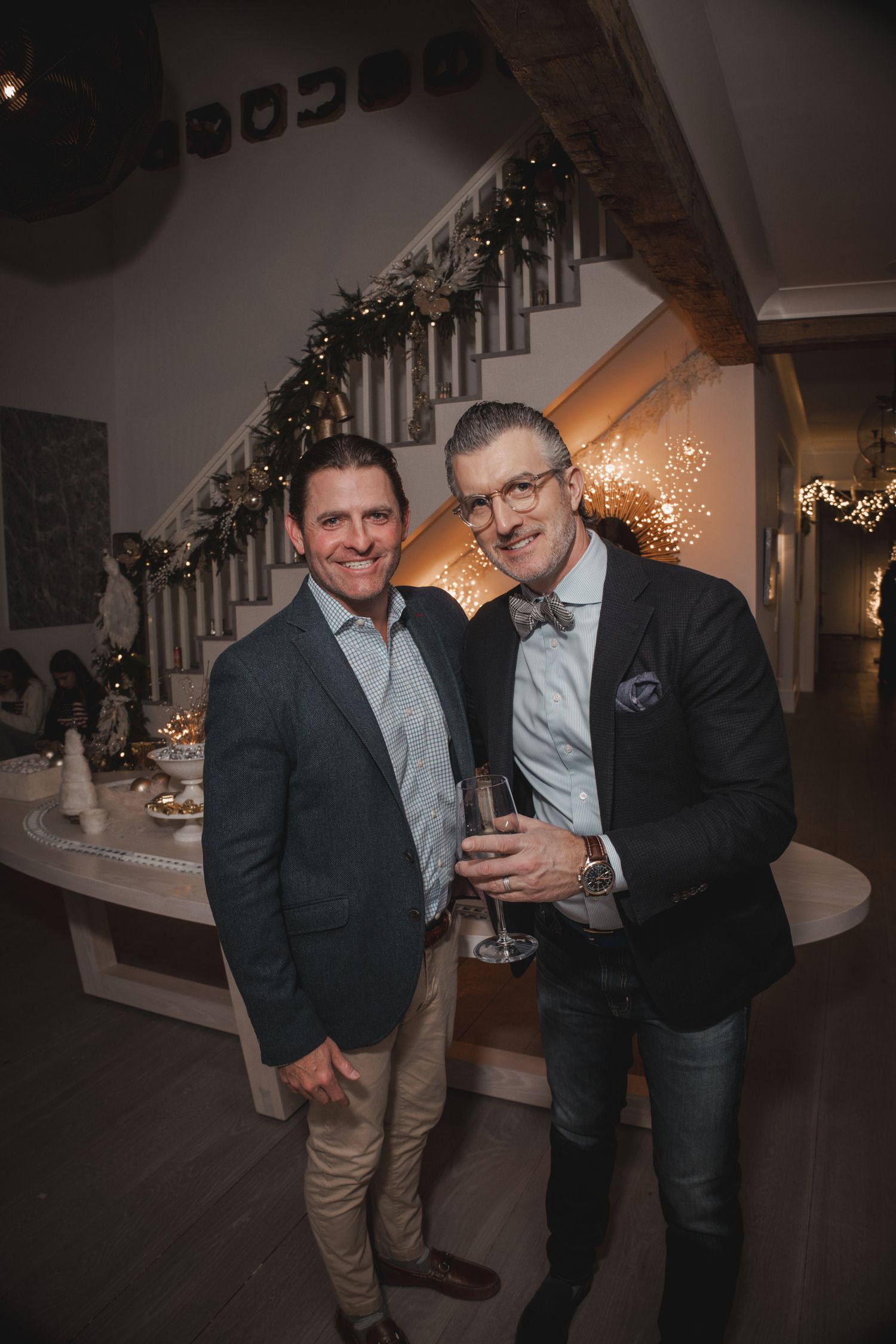 Two men smiling at a festive holiday party with garland-decorated staircase, one holding a wine glass.