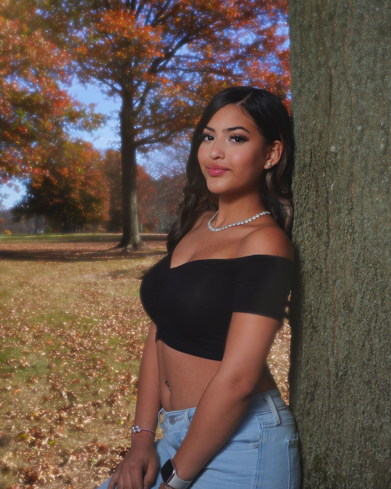 Woman in black crop top and denim poses against tree with autumn leaves in background.