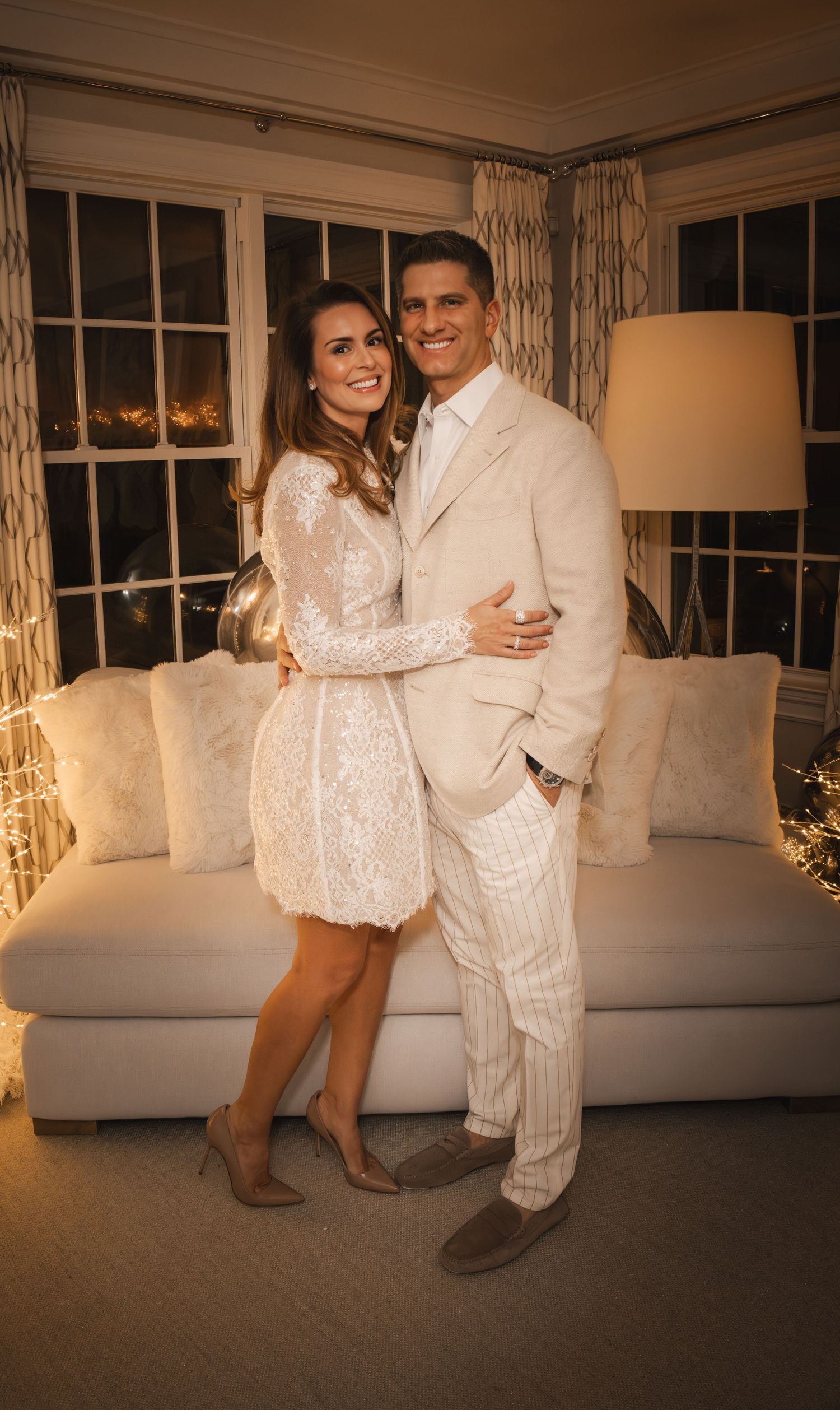 A couple in matching white outfits poses together in front of a couch in a warmly lit living room.