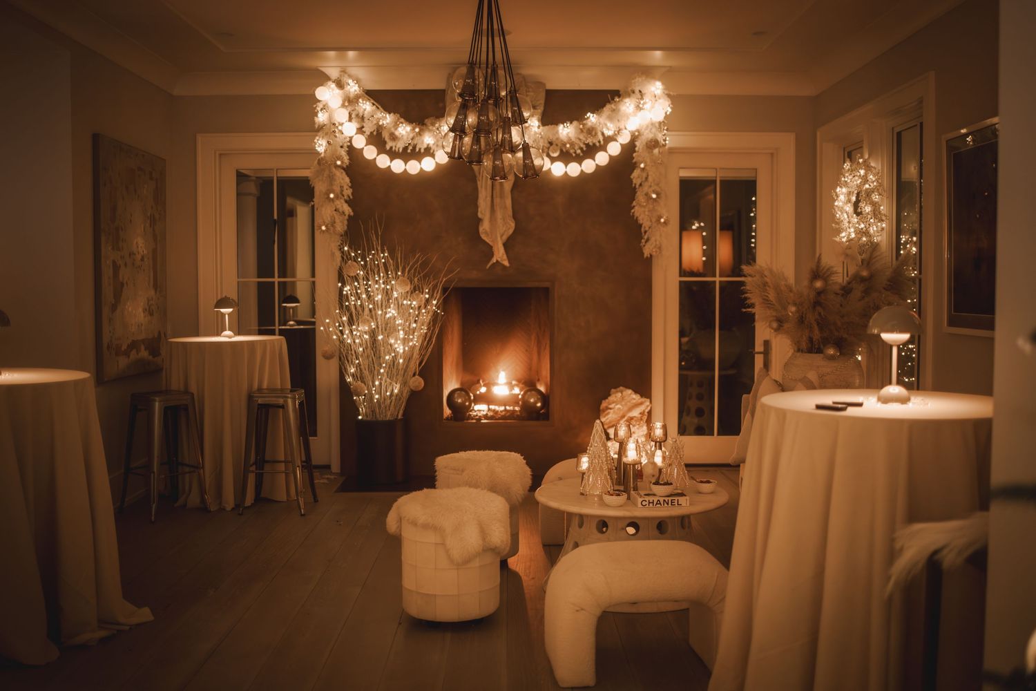 Cozy event space with warm fireplace, string lights, white lounge chairs, and elegant table settings in soft golden lighting.