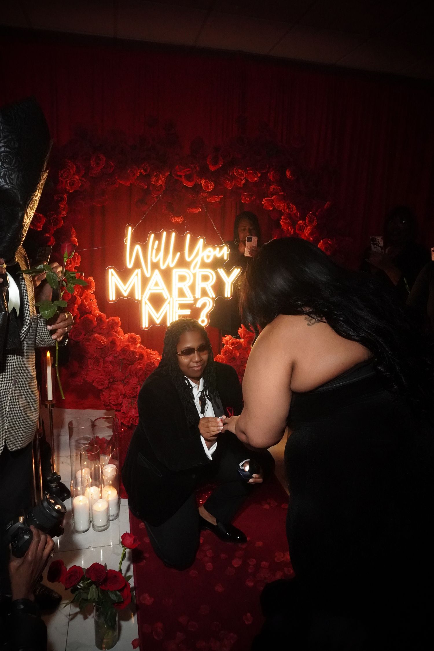 Marriage proposal scene with neon sign and red rose backdrop in romantic setting.