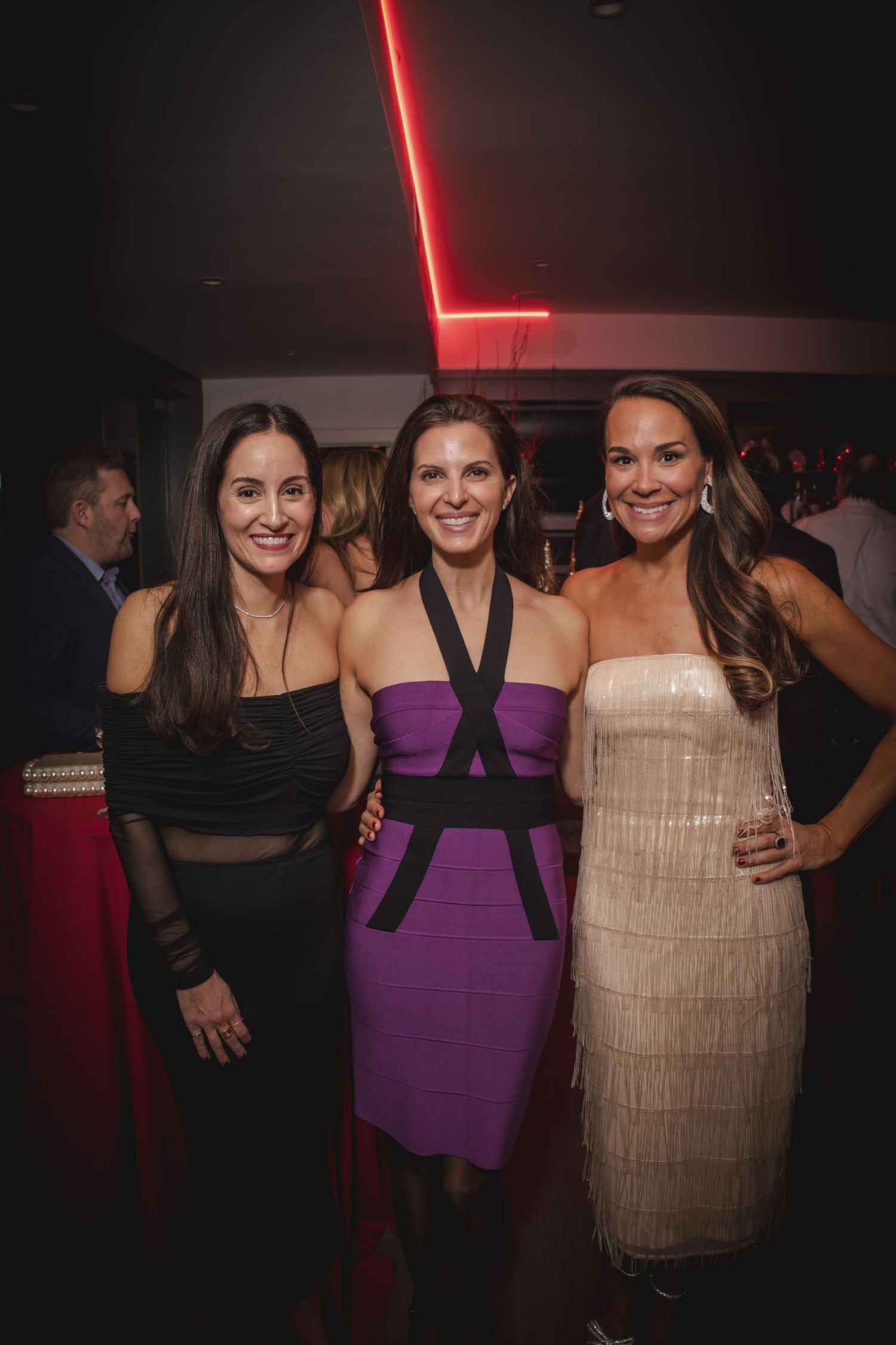 Three women pose together at a nightclub wearing black, purple and beige dresses in a dimly lit setting.
