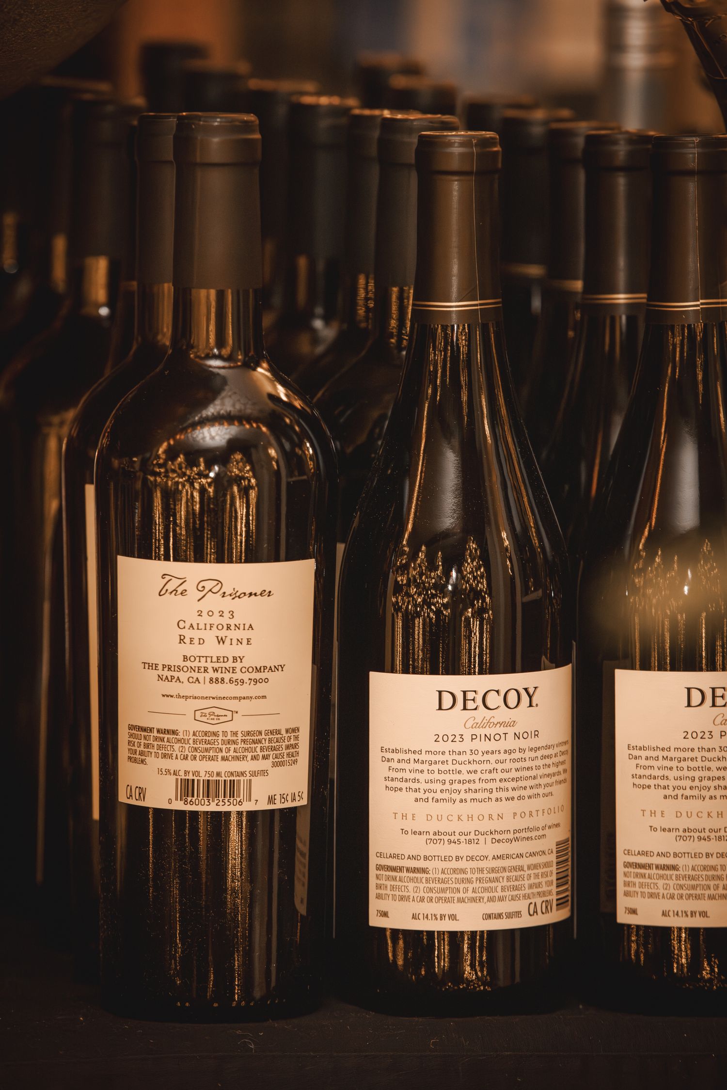 Bottles of Decoy 2019 Pinot Noir and The Prisoner red wine displayed on a shelf in warm lighting.