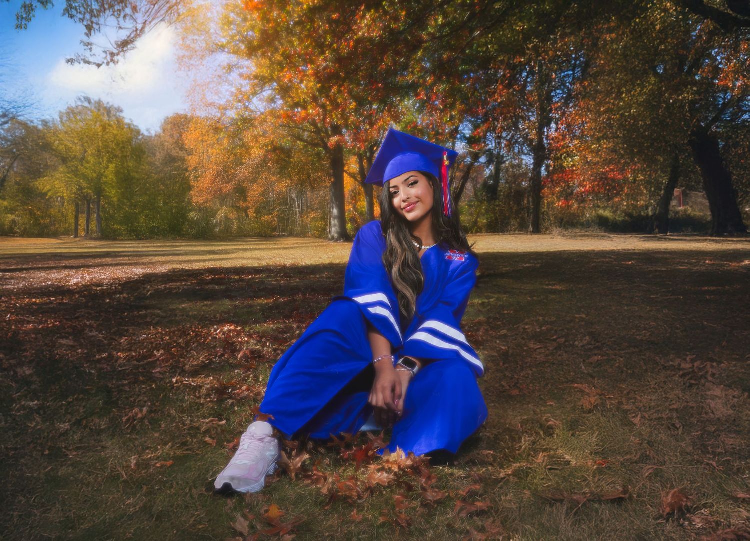 Graduate in blue cap and gown sits on fallen autumn leaves under trees.