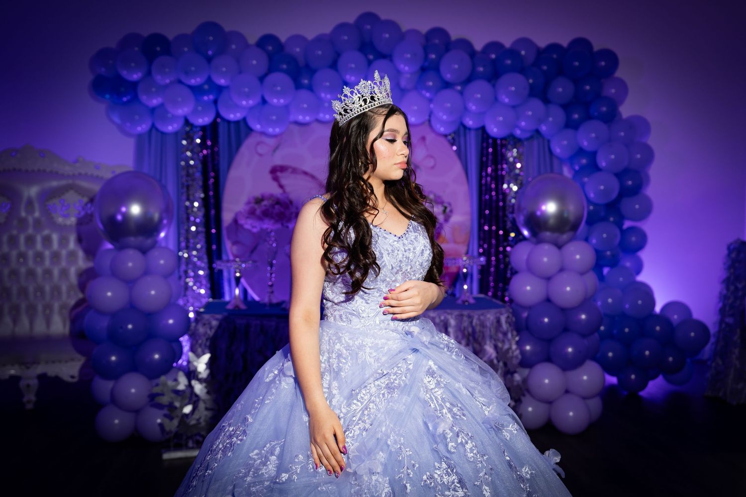 Celebratory portrait in lavender gown with crown against purple balloon backdrop.