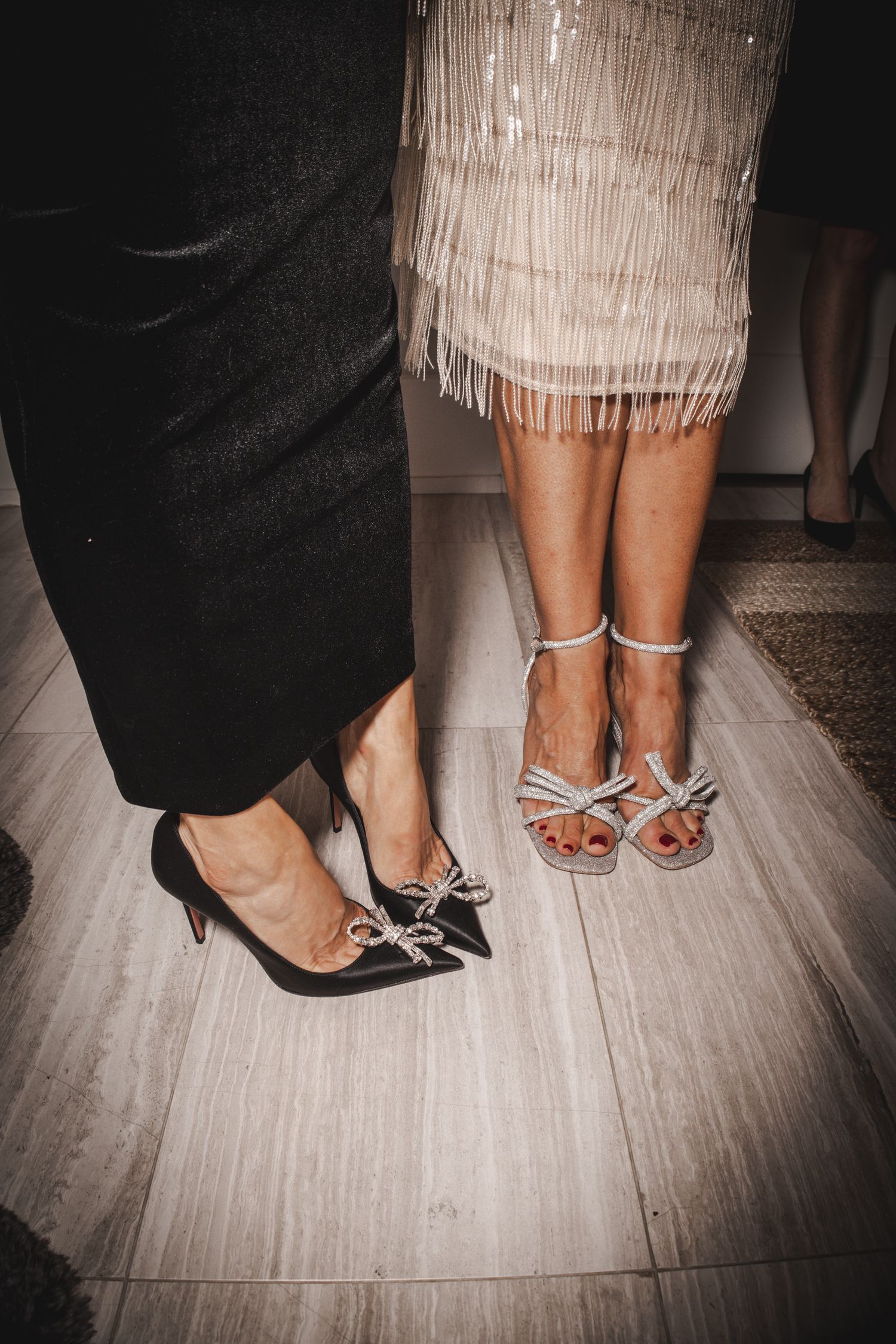 Two women standing side by side, one in black heels with bow detail, the other in strappy silver sandals with a fringed dress.