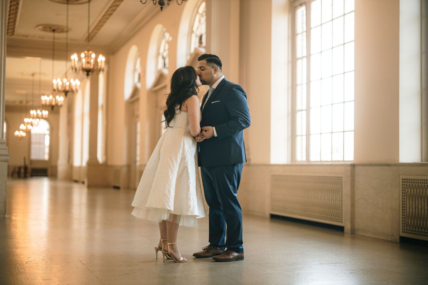 A couple shares a romantic moment in a bright ballroom with vintage chandeliers and arched windows.