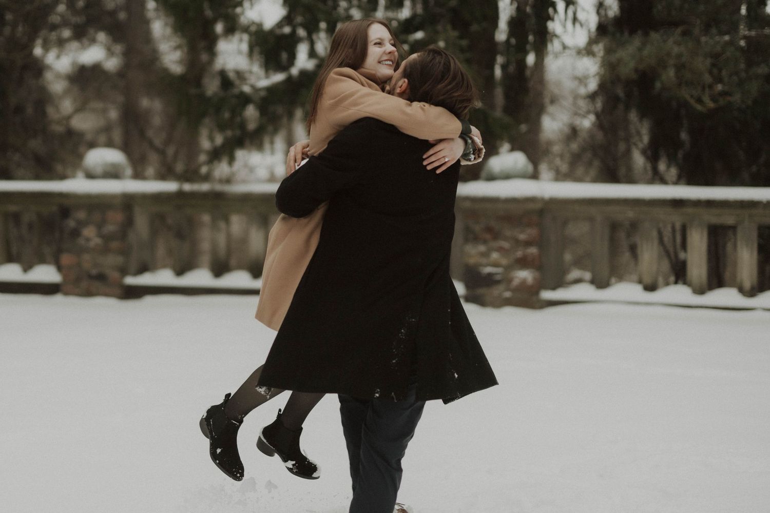 A couple embraces joyfully in a snowy winter scene with pine trees and a wooden railing in the background.