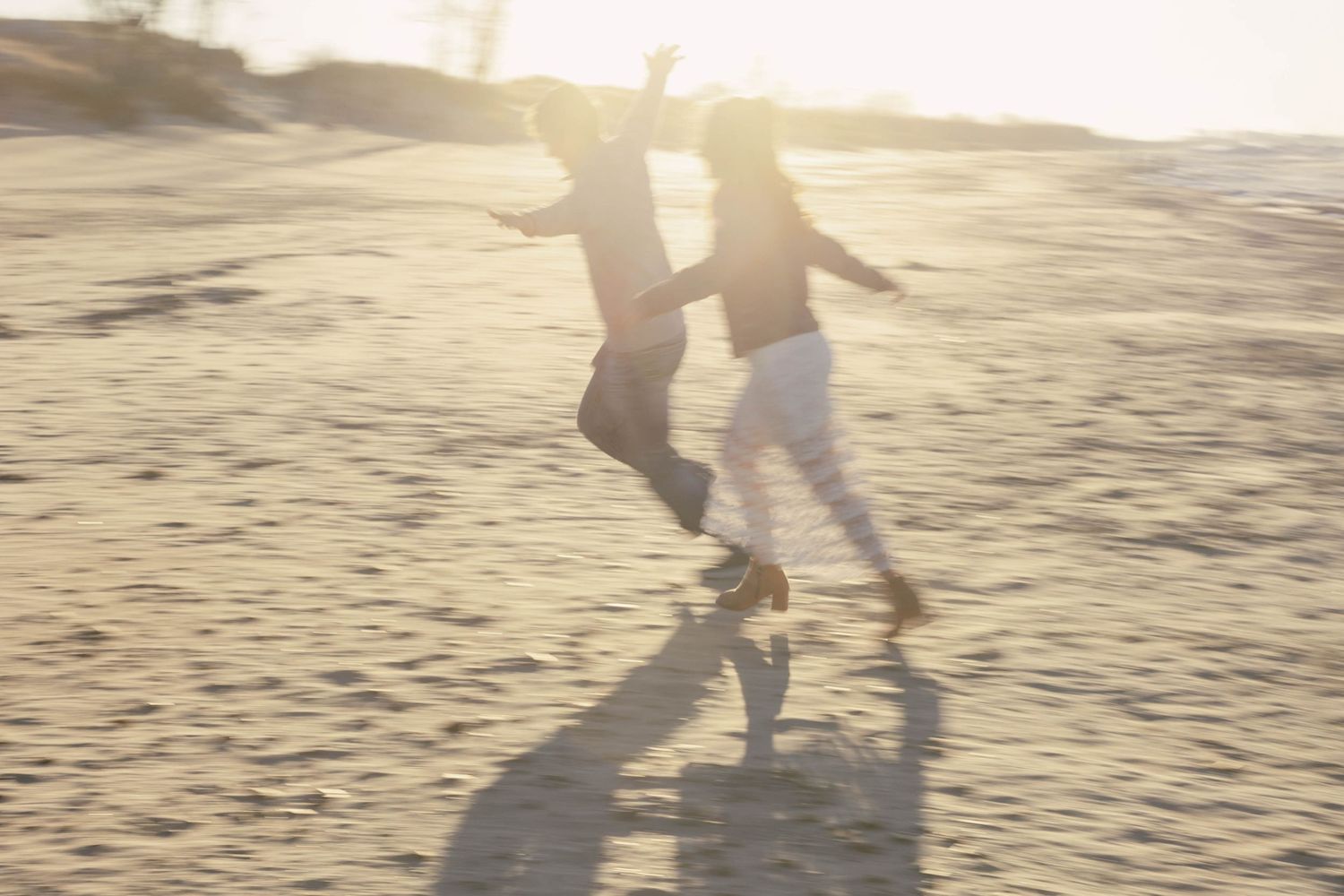 Two people walk together on a sandy beach at sunset casting long shadows on the sand.