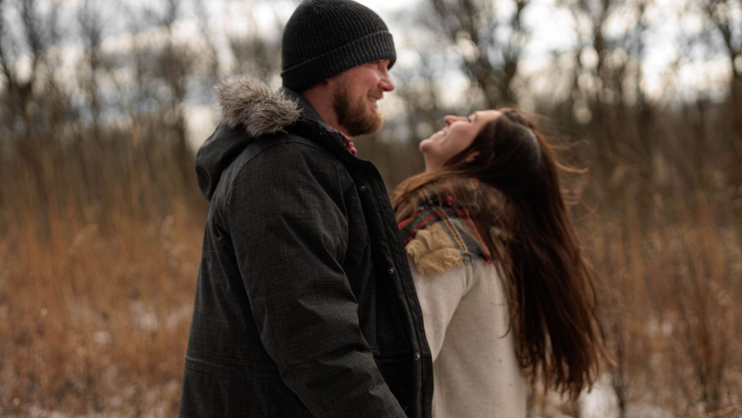 A couple embraces in a wintry field with dried grass and bare trees in the background during golden hour.