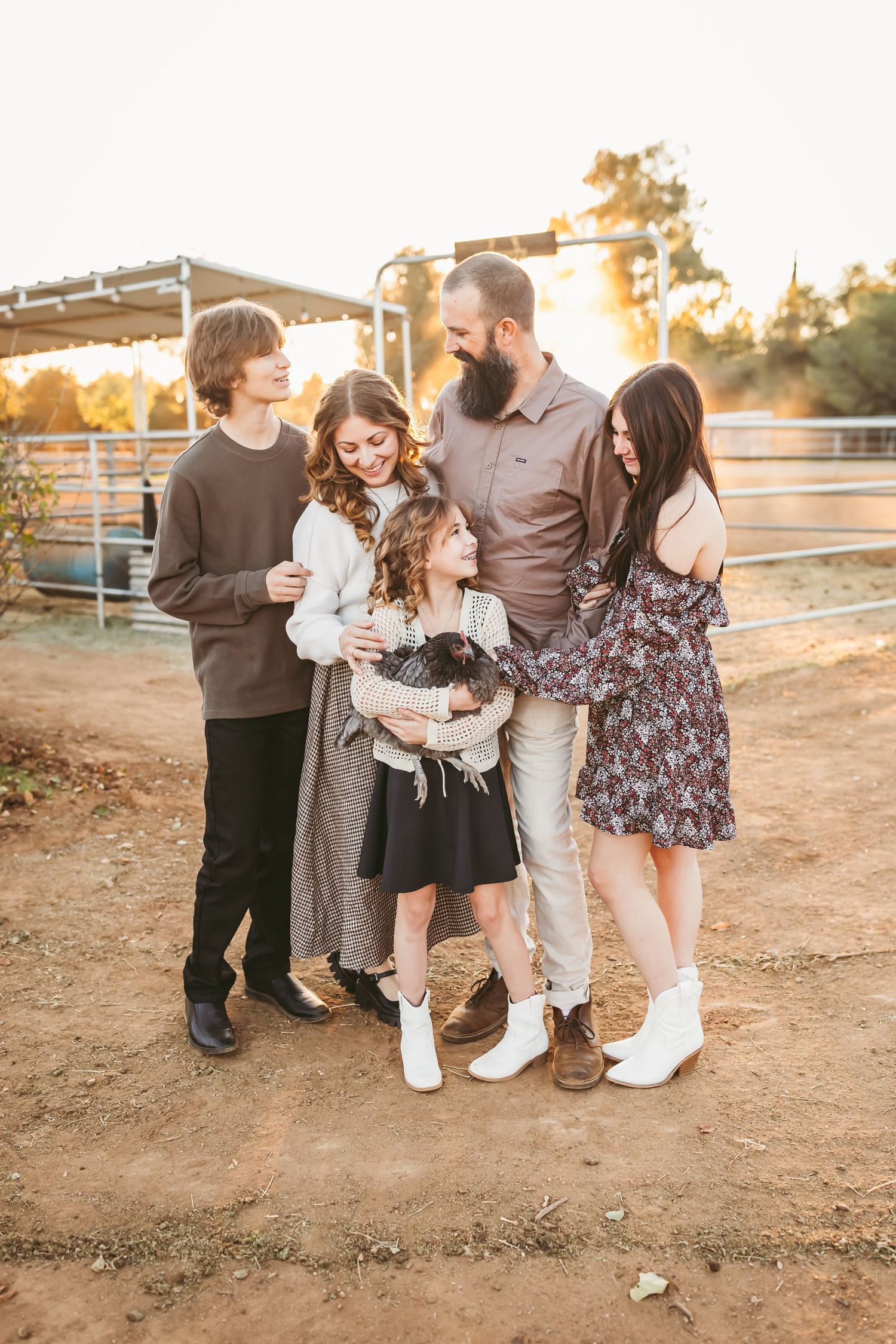 A family group laughs together while standing outdoors at sunset on a dirt path near white fencing.