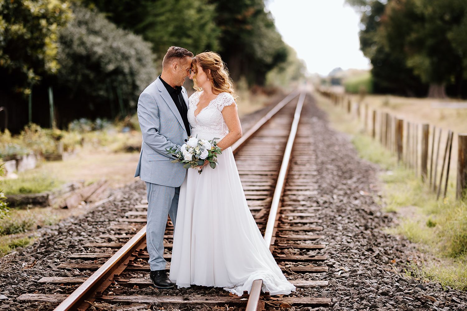 A romantic wedding couple shares an intimate moment while standing on train tracks in soft natural lighting.