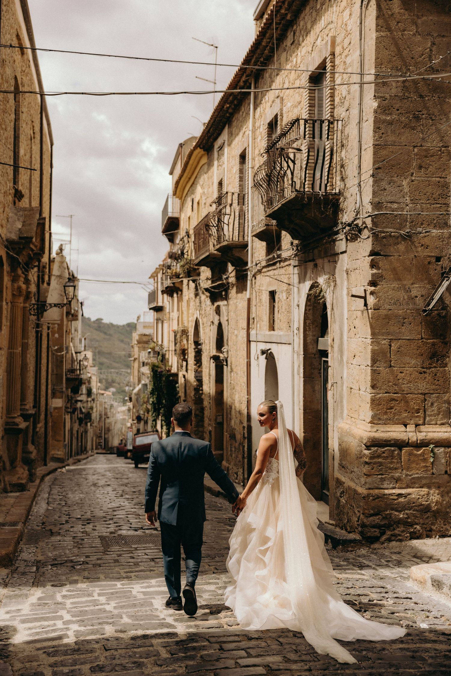 A couple strolls down a charming cobblestone street in an old European town with historic stone buildings.