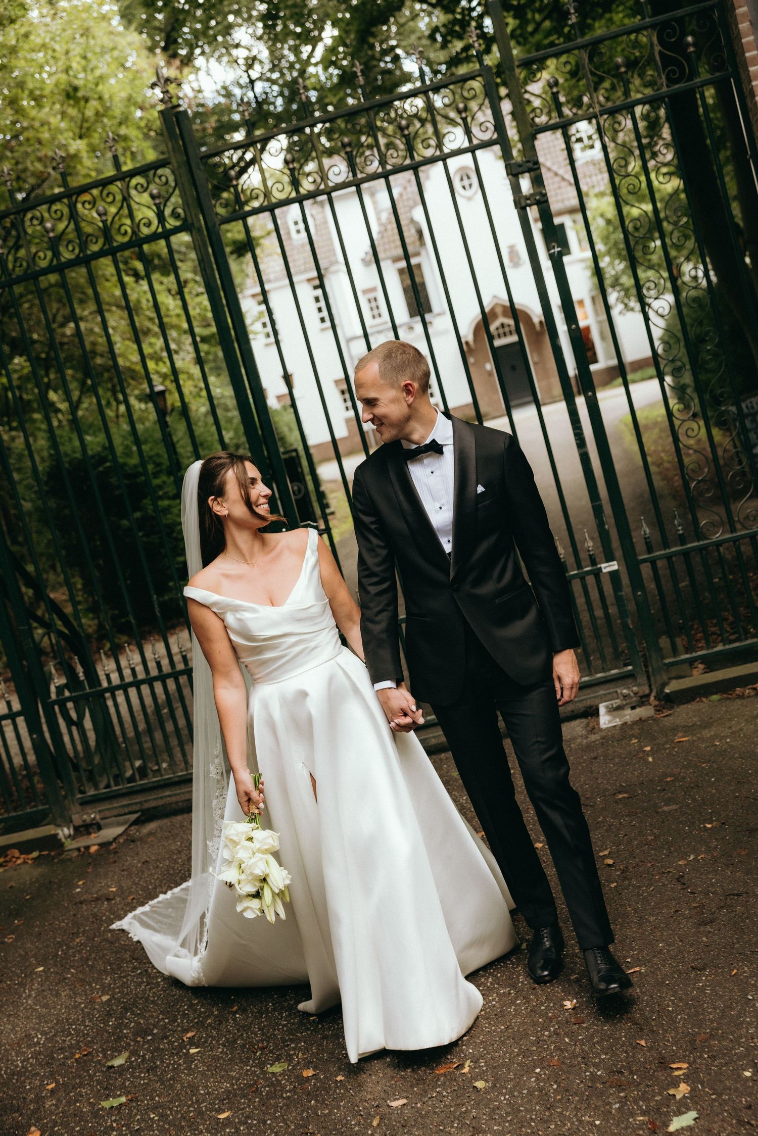 Bride in white gown and groom in black tuxedo hold hands smiling before ornate iron gates outdoors.