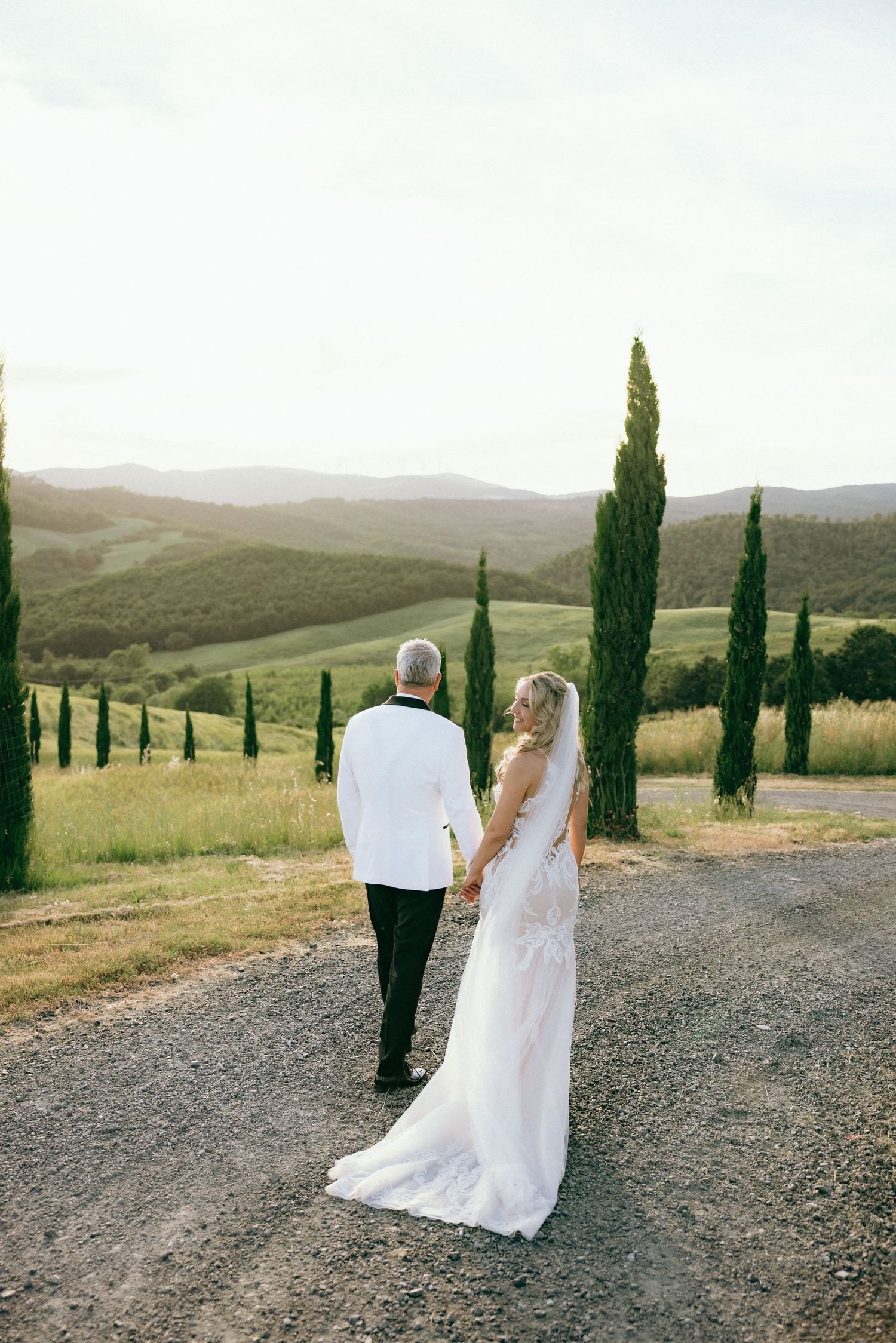 A couple strolls down a winding country road with cypress trees against a golden Tuscan landscape.