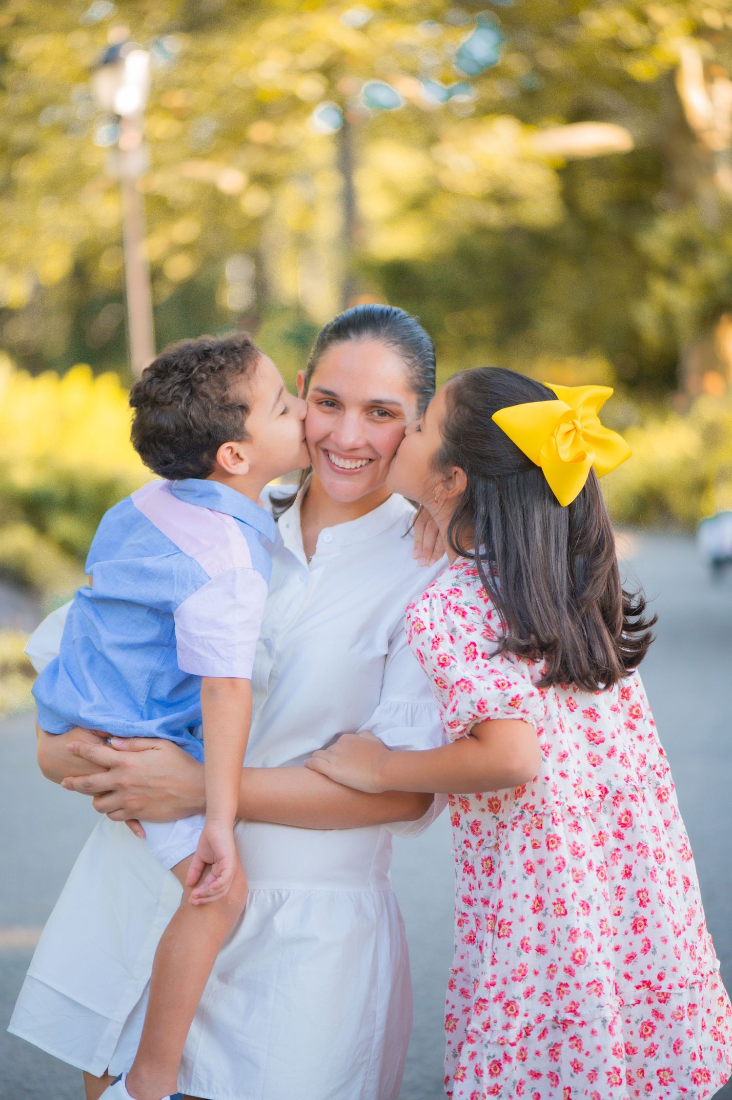 A smiling mother in a white dress holds her young son while he and a girl with a yellow bow kiss her cheeks outdoors.