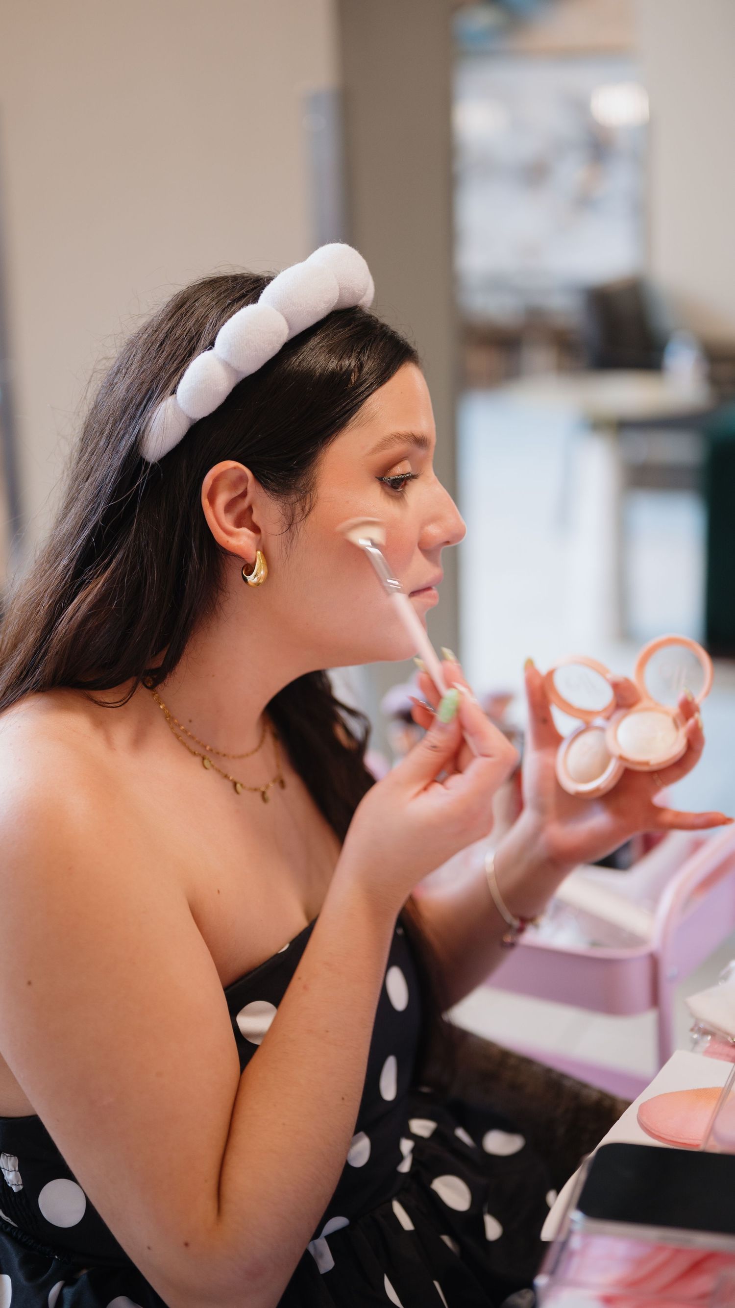 Woman in a polka dot top and white headband applies blush with a brush while holding a compact of pink and peach makeup shades.