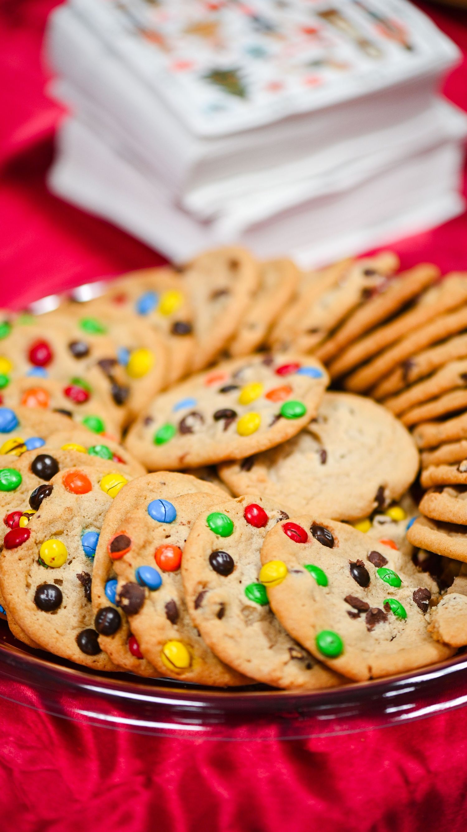 Platter of colorful candy-studded chocolate chip cookies on a red tablecloth with stacked party napkins behind.