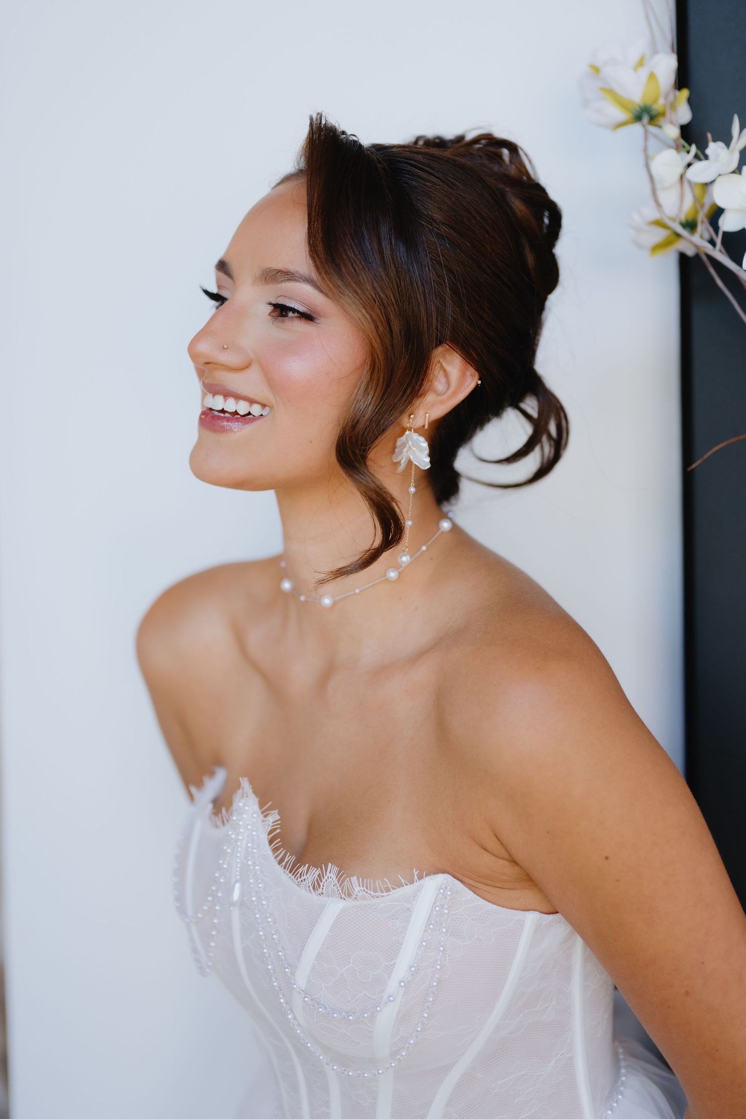 Smiling bride in a white strapless lace corset gown with an updo, pearl necklace and floral drop earrings, posed beside white blossoms.