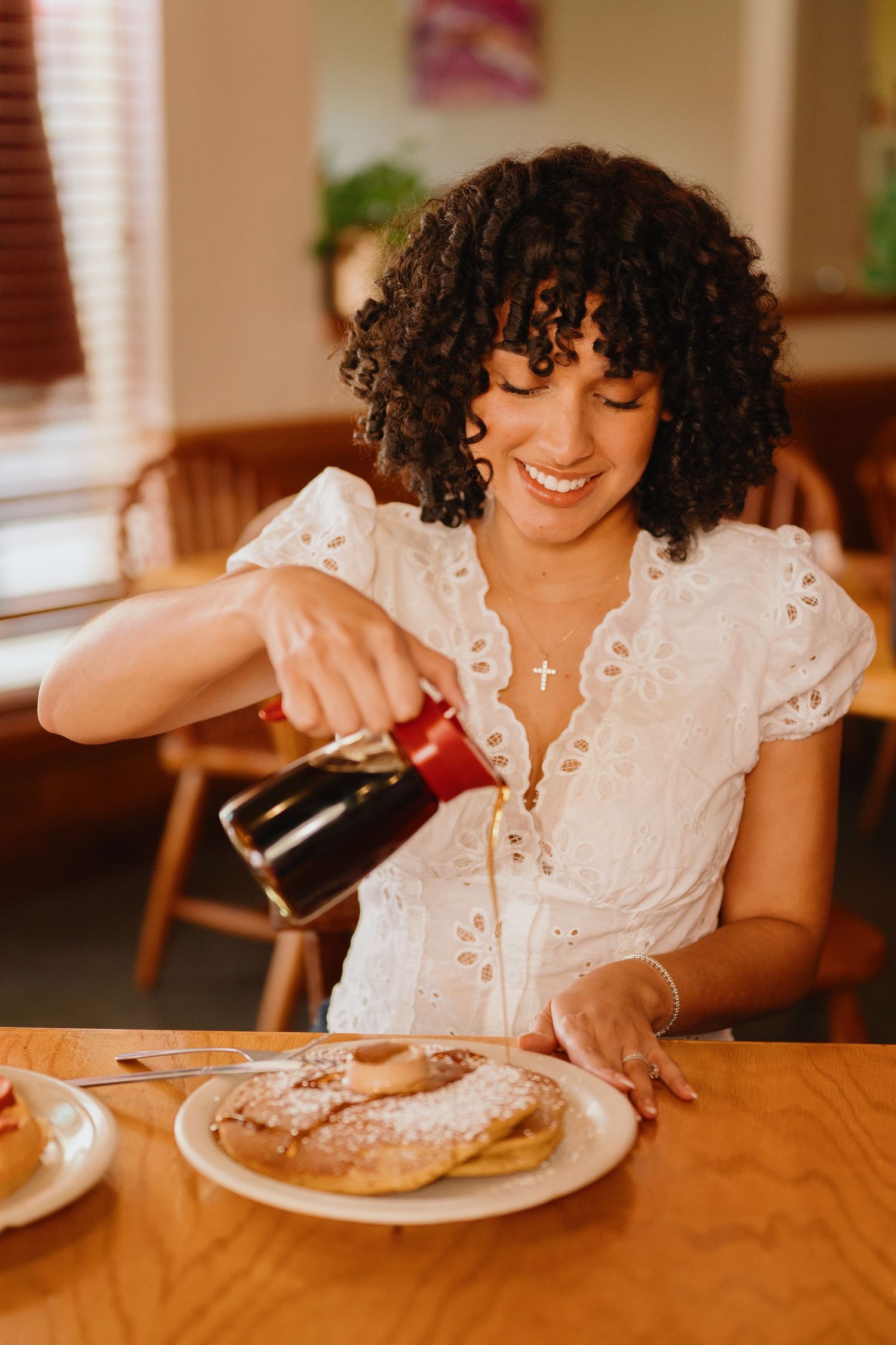 Smiling woman with curly hair pours syrup over a stack of fluffy pancakes at a wooden restaurant table.