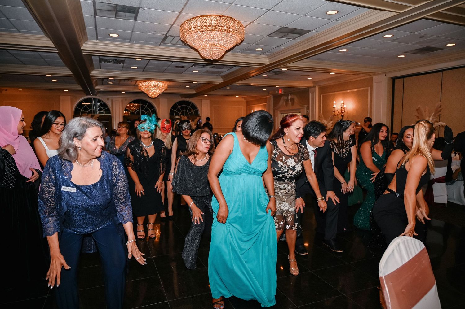 Guests in evening dresses dance together on a ballroom floor beneath a chandelier at a festive gala.