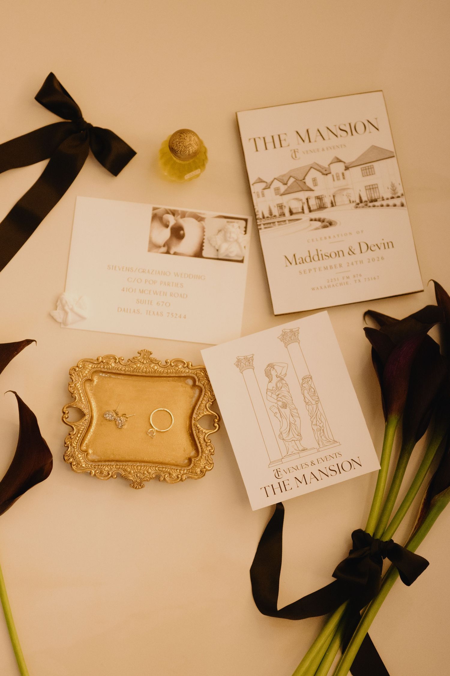 Wedding stationery flat lay with invitations, gold ring tray, black ribbons, and dark calla lilies from The Mansion venue.