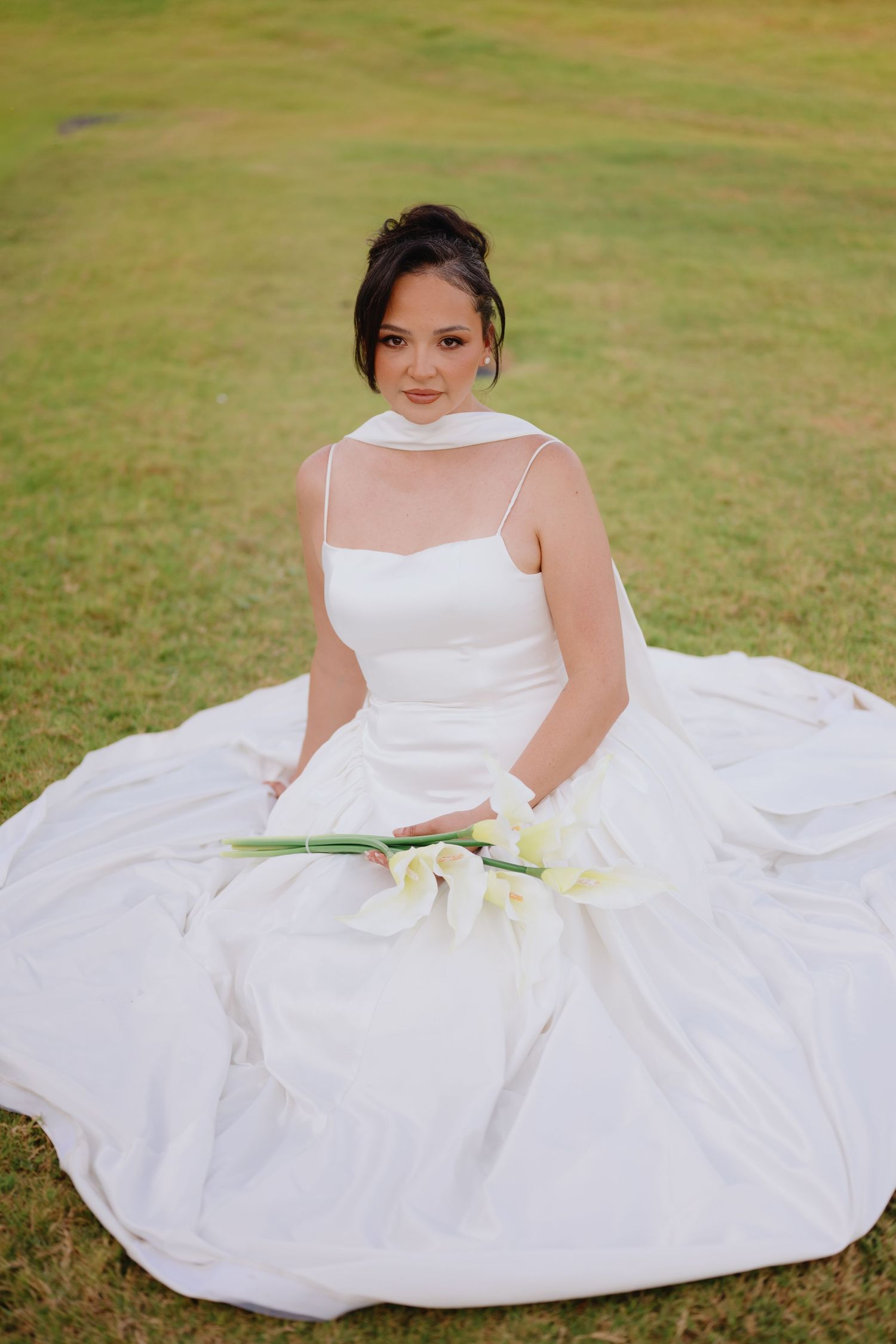 Bride in a satin gown with a sleek halter wrap sits on a grassy lawn holding a bouquet of white calla lilies.