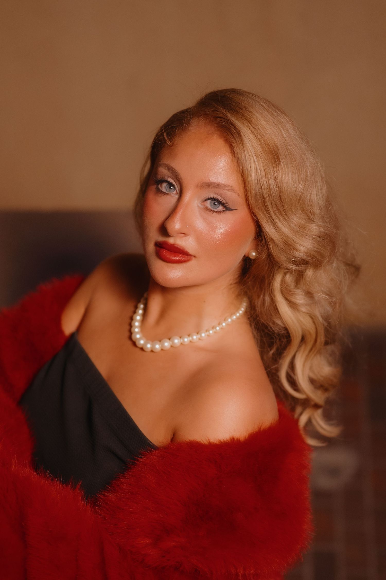 Woman with wavy blonde hair, red lipstick and a pearl necklace poses in a black dress with a red faux fur wrap.